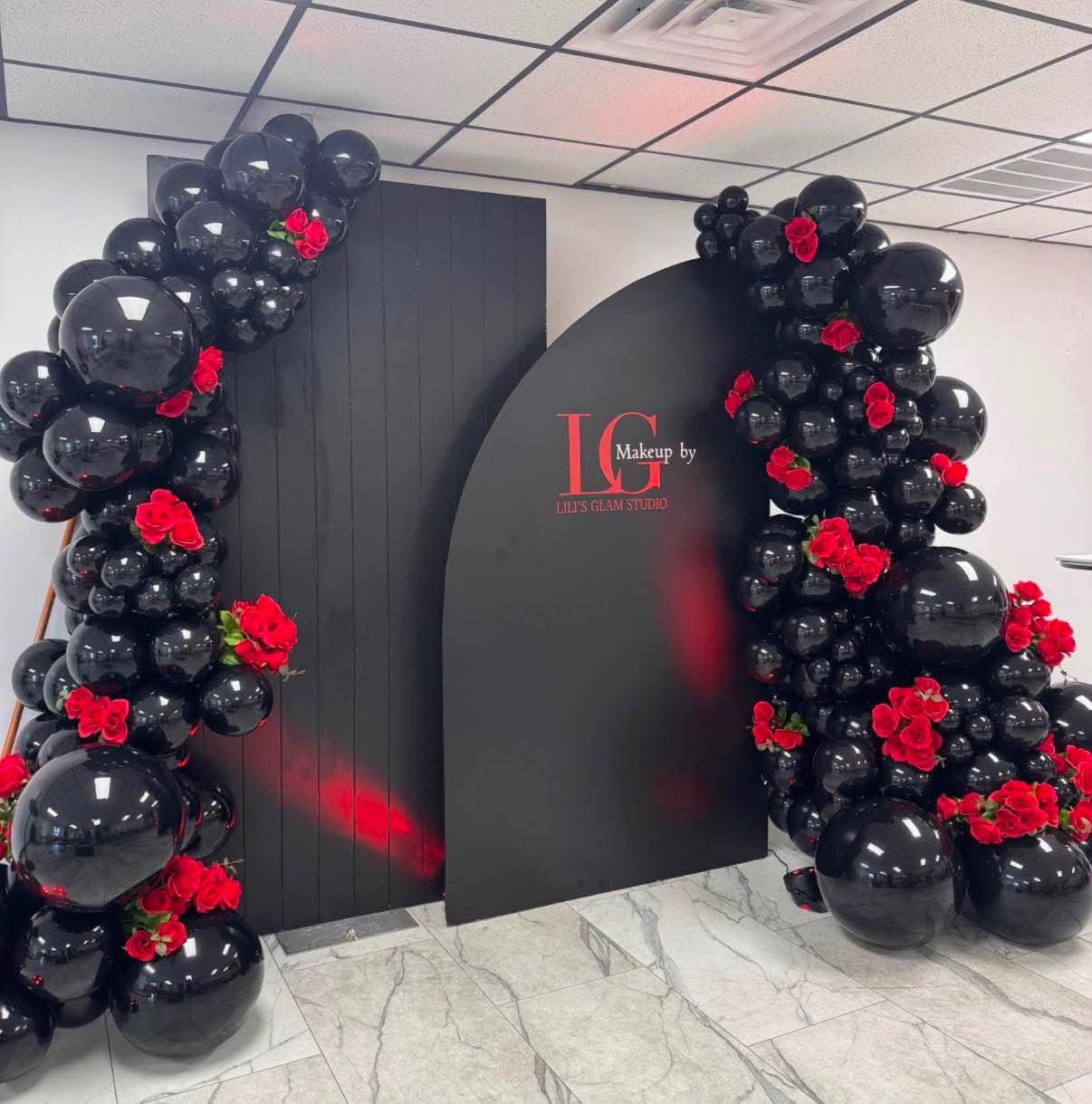 Decorative black balloon arch with red flowers surrounds a black wall with red text, in an indoor setting.