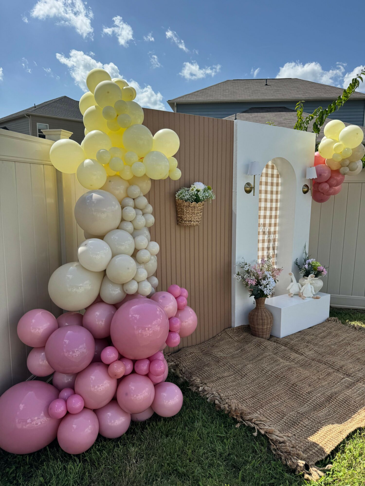 Decorative setup with balloons, a small table, and flowers outdoors, with a blue sky and clouds in the background.
