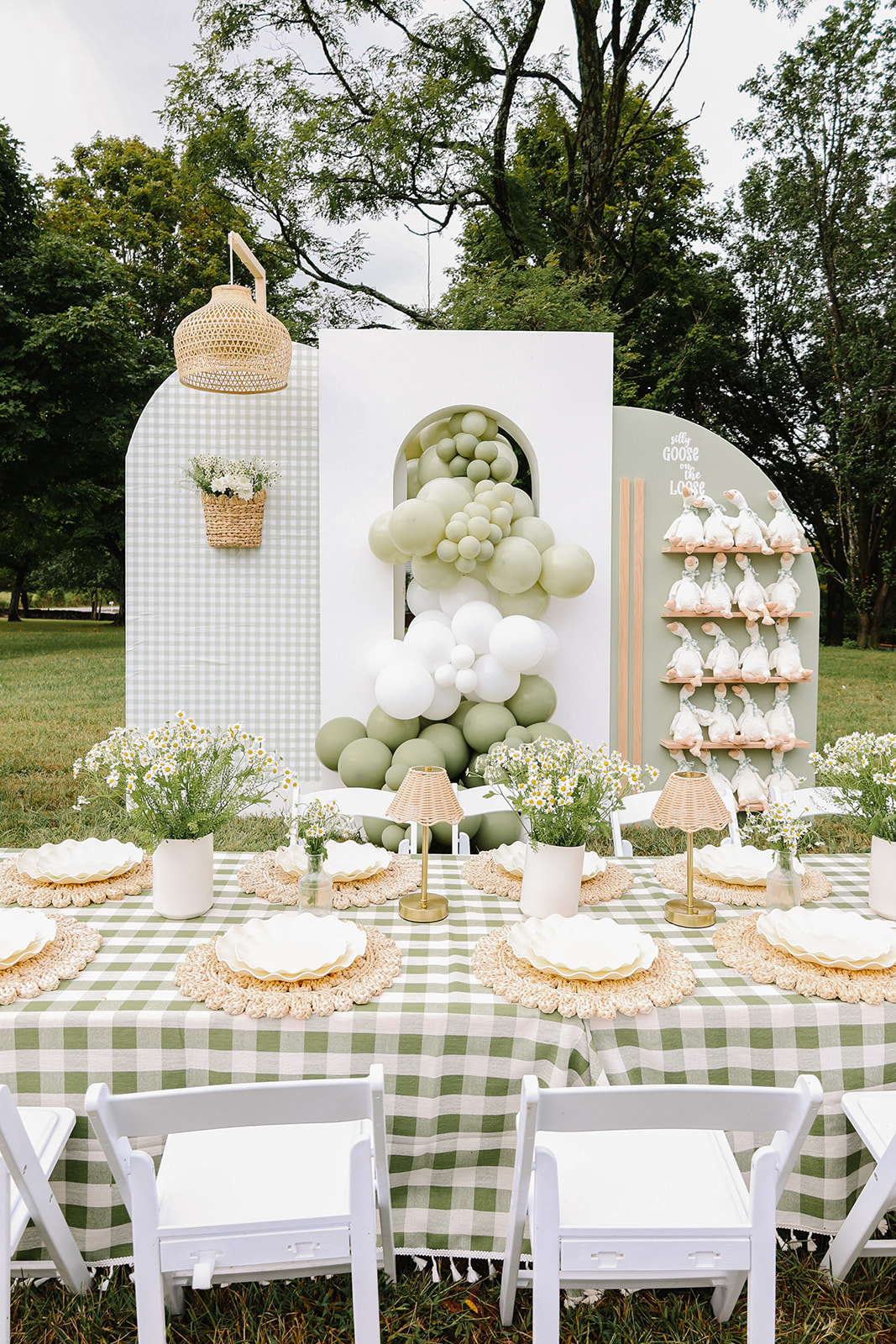 Outdoor table with green checkered tablecloth, white chairs, floral centerpieces, and dessert plates, set in a park with trees.