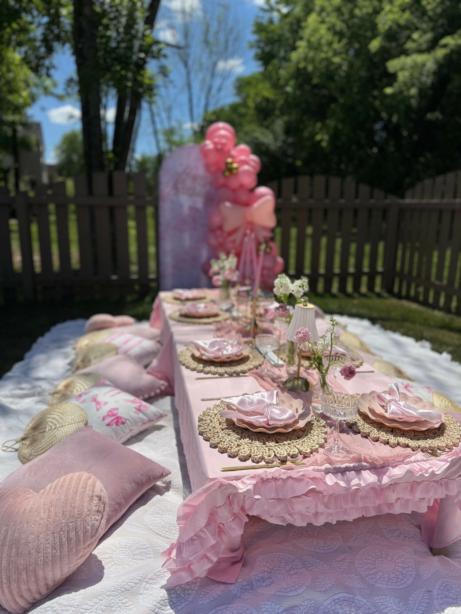 Outdoor table set with pink decorations, balloons, and floral arrangements, surrounded by trees and a wooden fence.