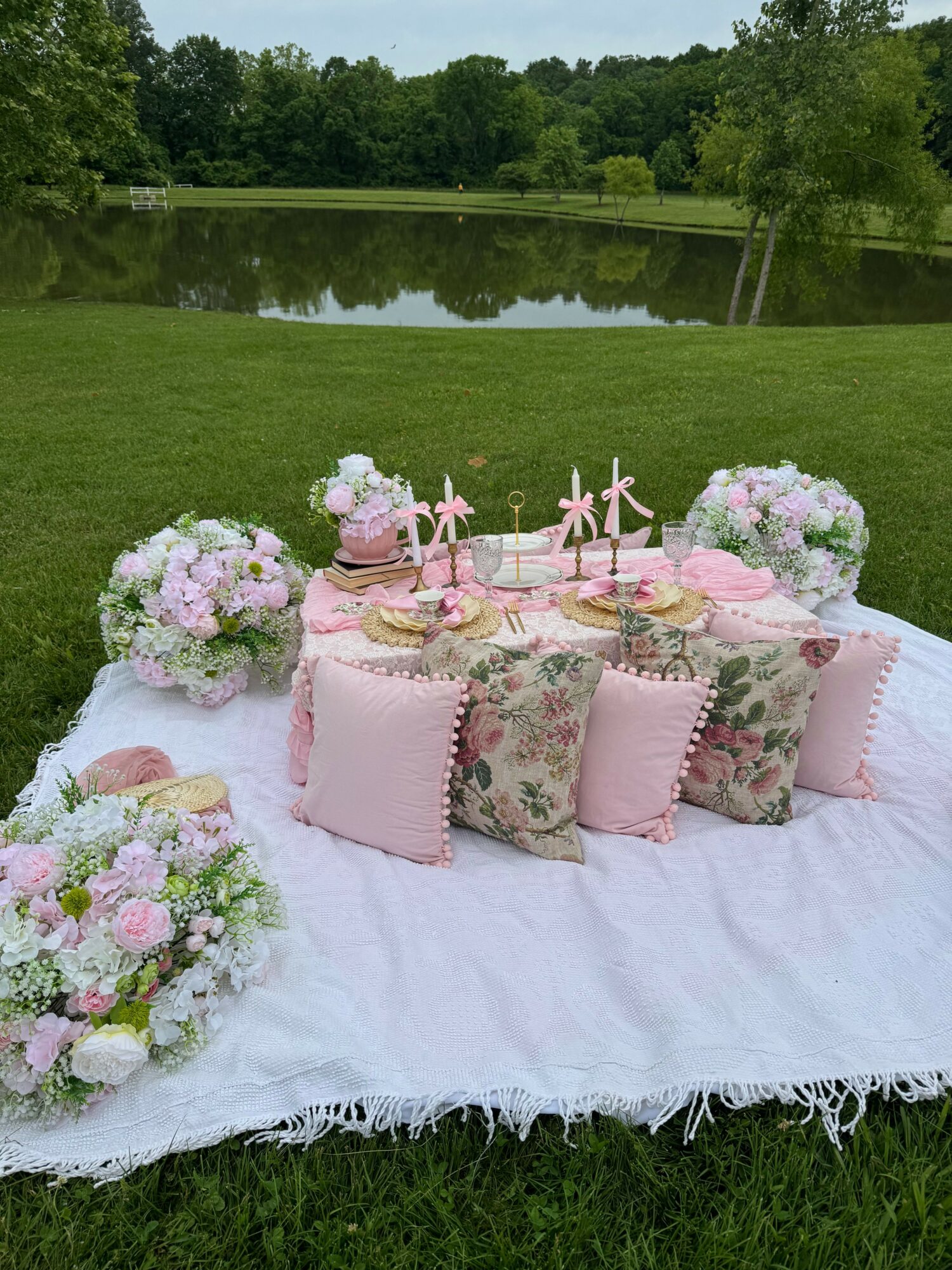 Outdoor table set with pink and floral tableware, surrounded by pink flowers on a white cloth, on grass.