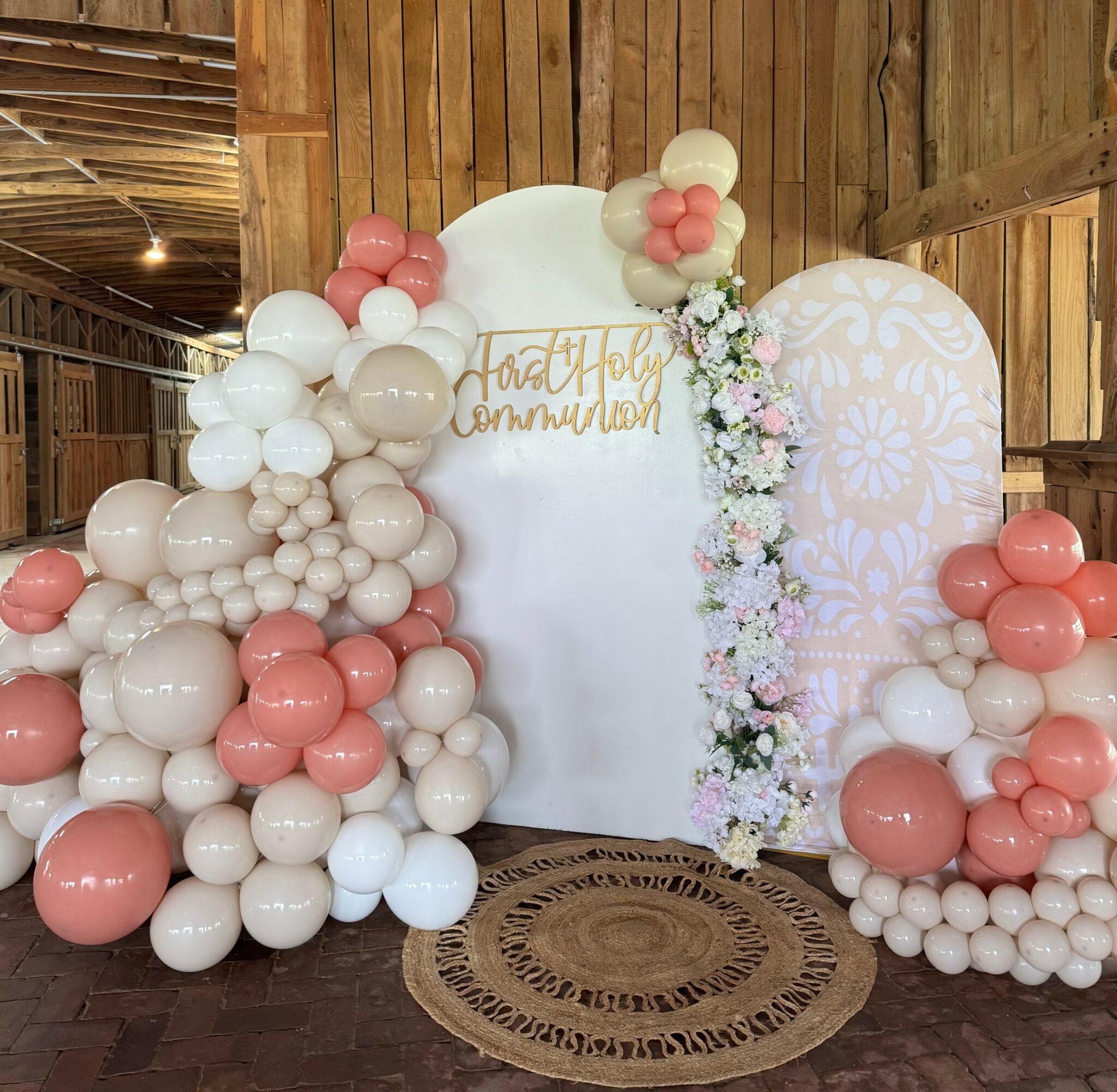Decorative balloon arrangement with pink, white, and beige balloons, floral garland, and a white backdrop with gold writing, in a wooden setting.