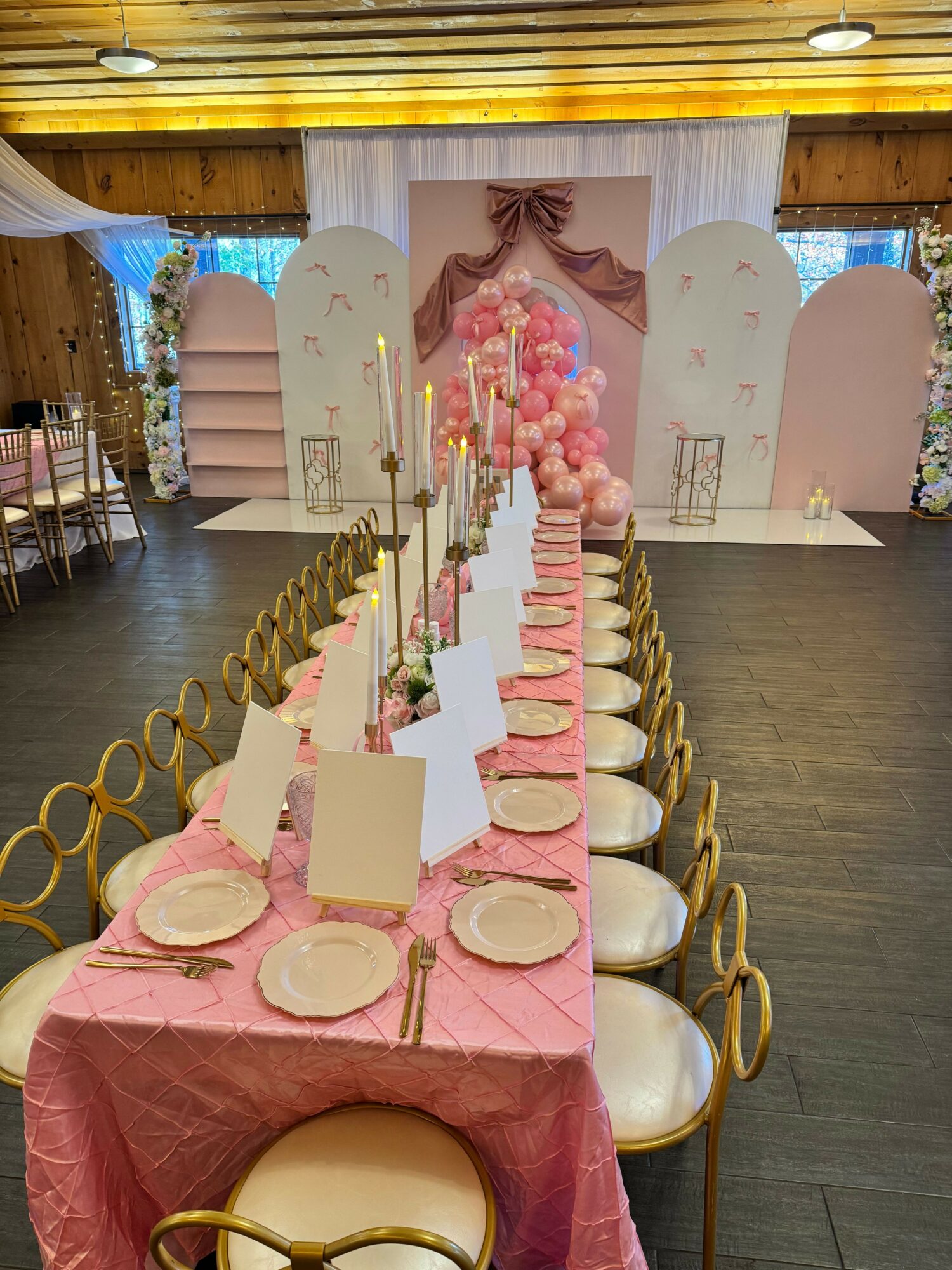 Long table set with pink tablecloth, gold chairs, plates, and napkins, decorated for a celebration with pink and gold theme.