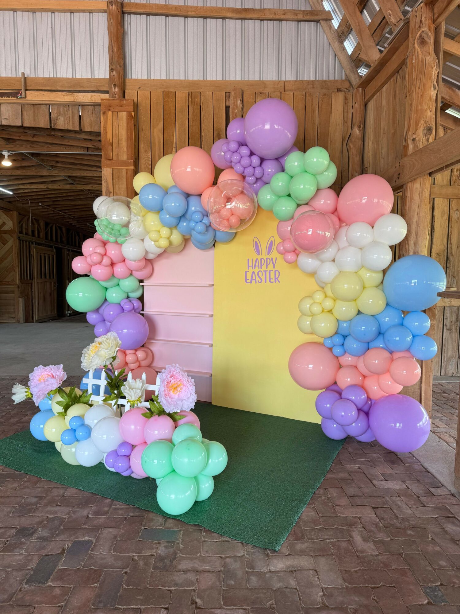 Colorful balloon arch with pink, purple, blue, green, and yellow balloons over a yellow backdrop with text, on a green carpet.