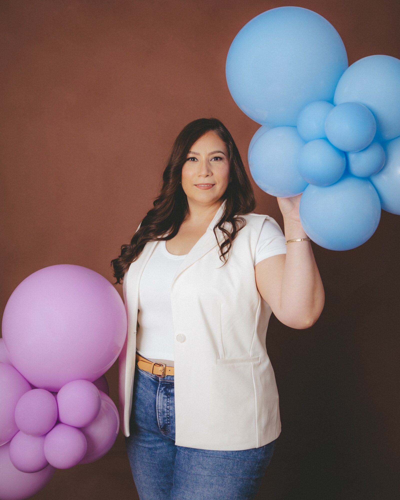 Woman holding large blue balloon cluster, standing against brown background, wearing white vest and jeans, smiling.