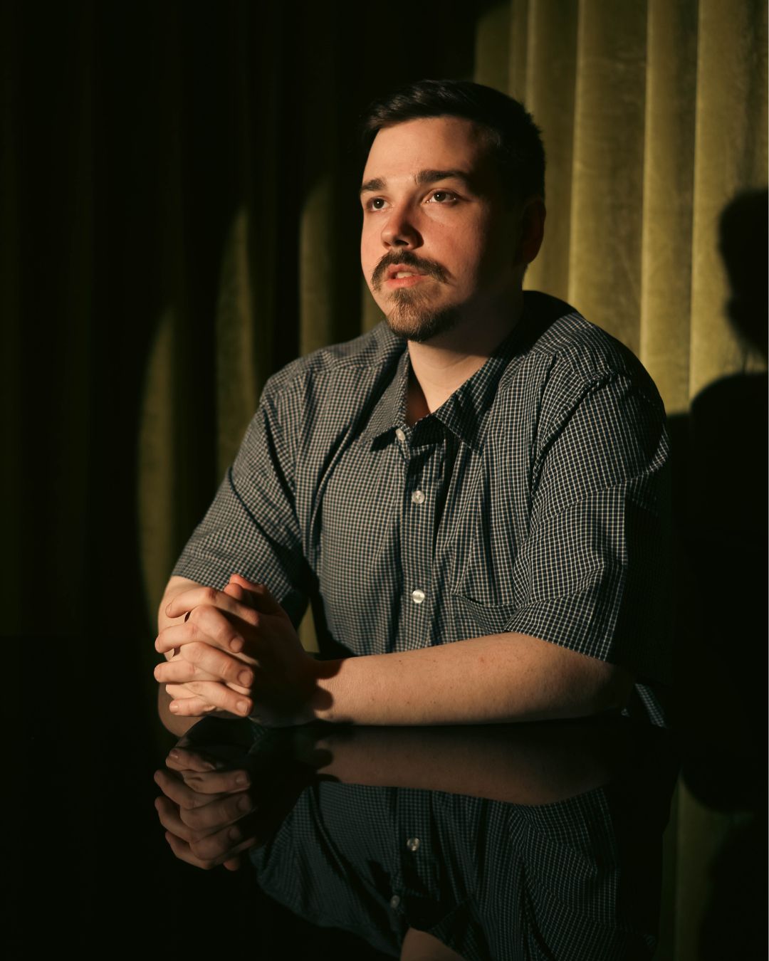 Man with dark hair and beard, wearing a dark striped shirt, sitting with hands clasped, in front of yellow curtains.