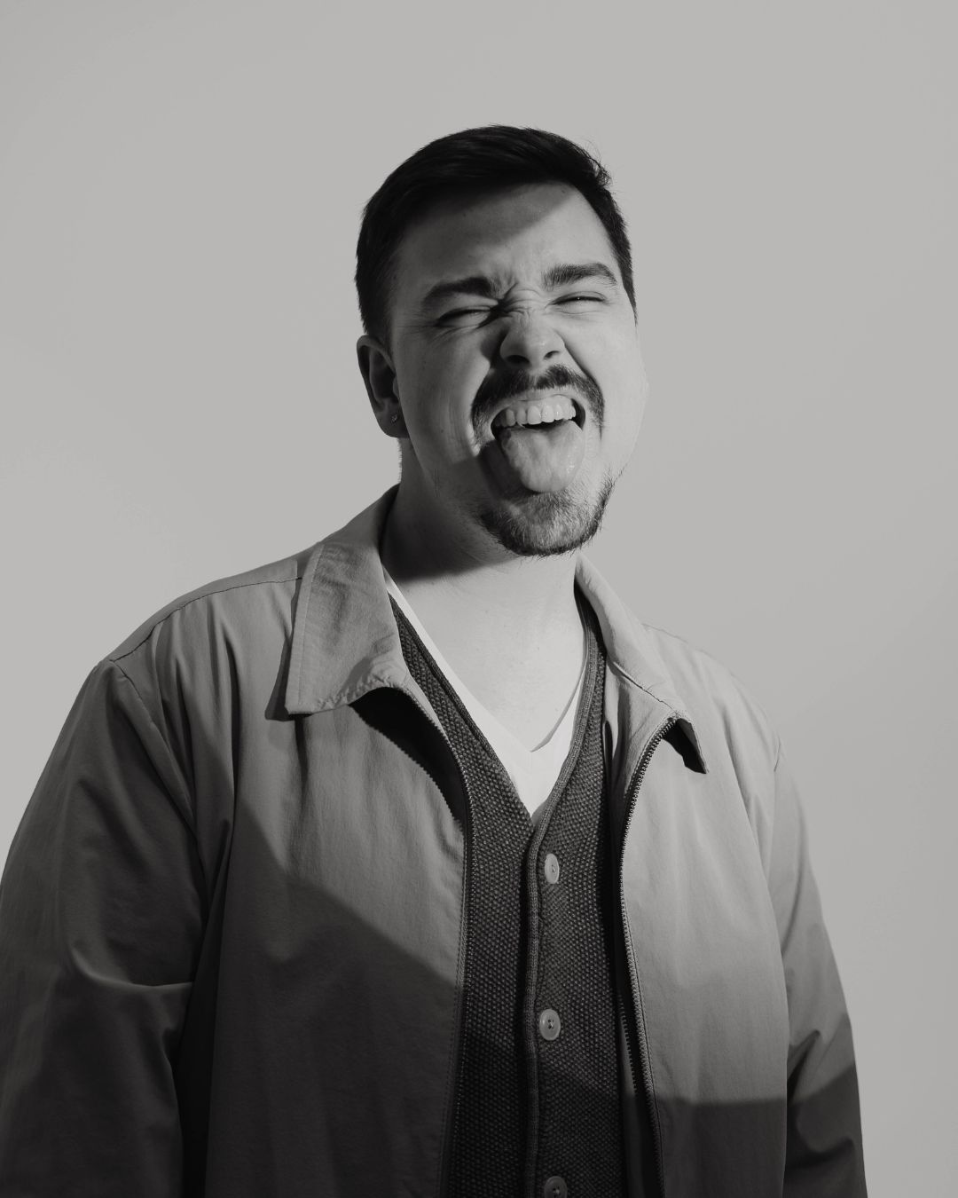 Man with short dark hair making a playful face, sticking out tongue, wearing a jacket and shirt, against plain background.