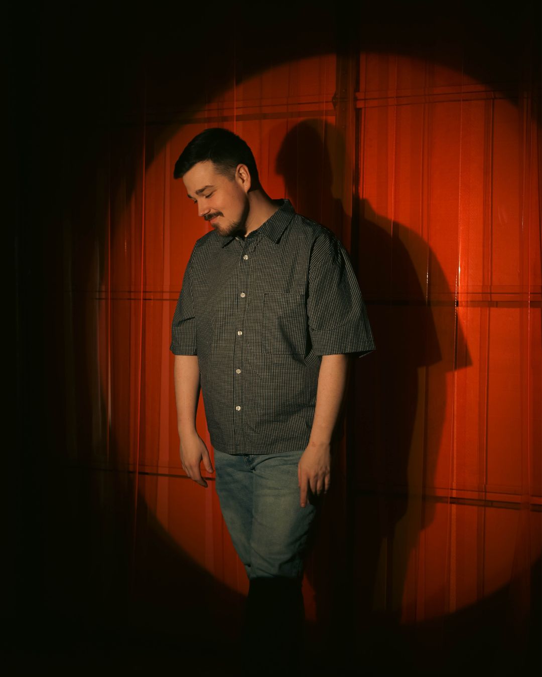 Young man standing on stage with a red curtain background, looking down, shadow cast behind him, spotlight focused on him.
