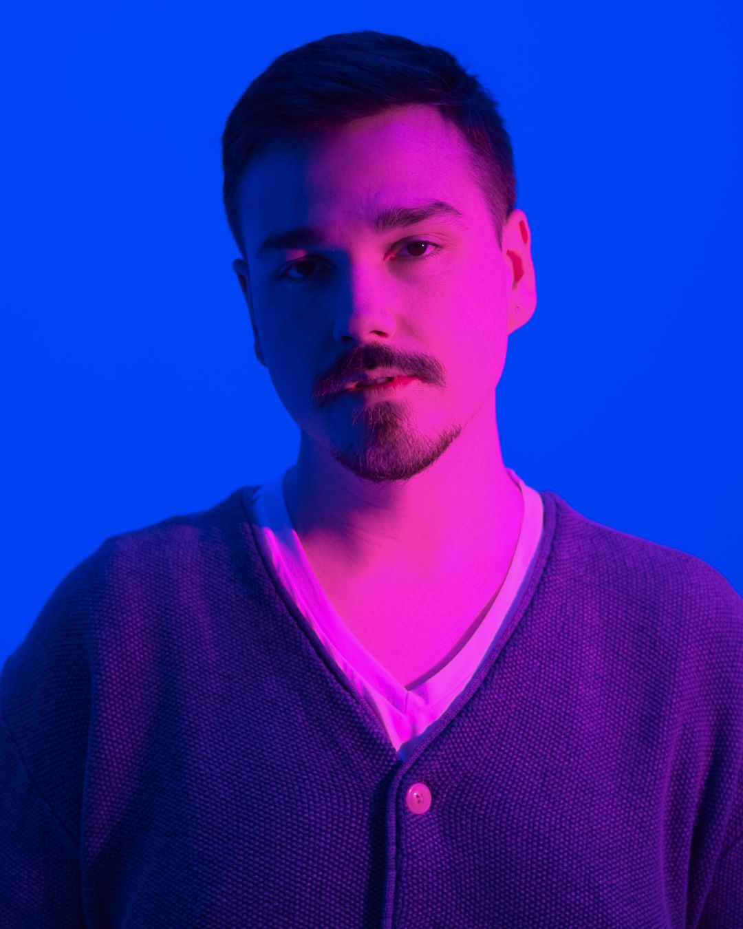Portrait of a man with short dark hair and facial hair, wearing a V-neck shirt and cardigan, against a blue background.