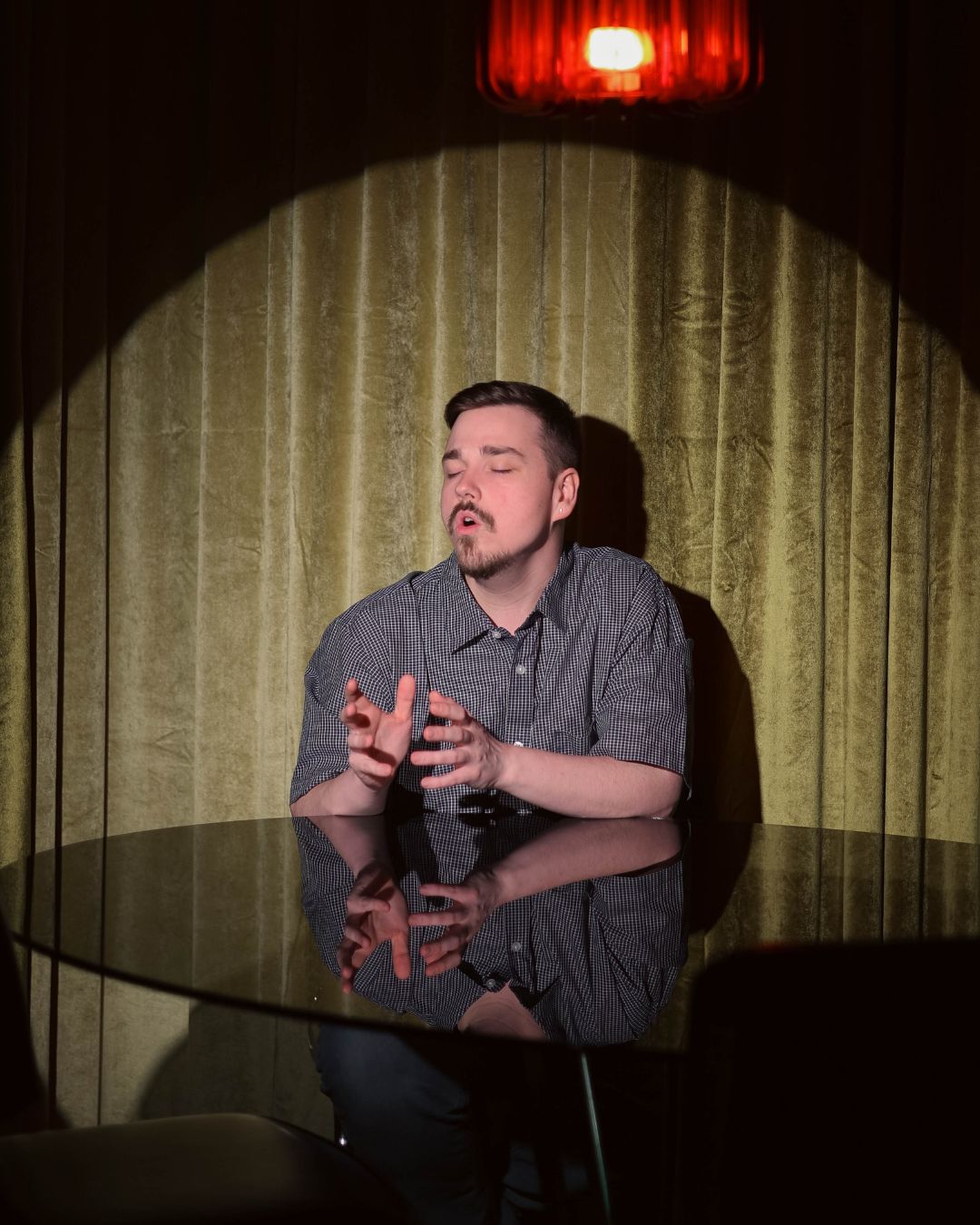 Man with closed eyes and hands raised, sitting at a reflective table in front of a green curtain, illuminated by a red light.