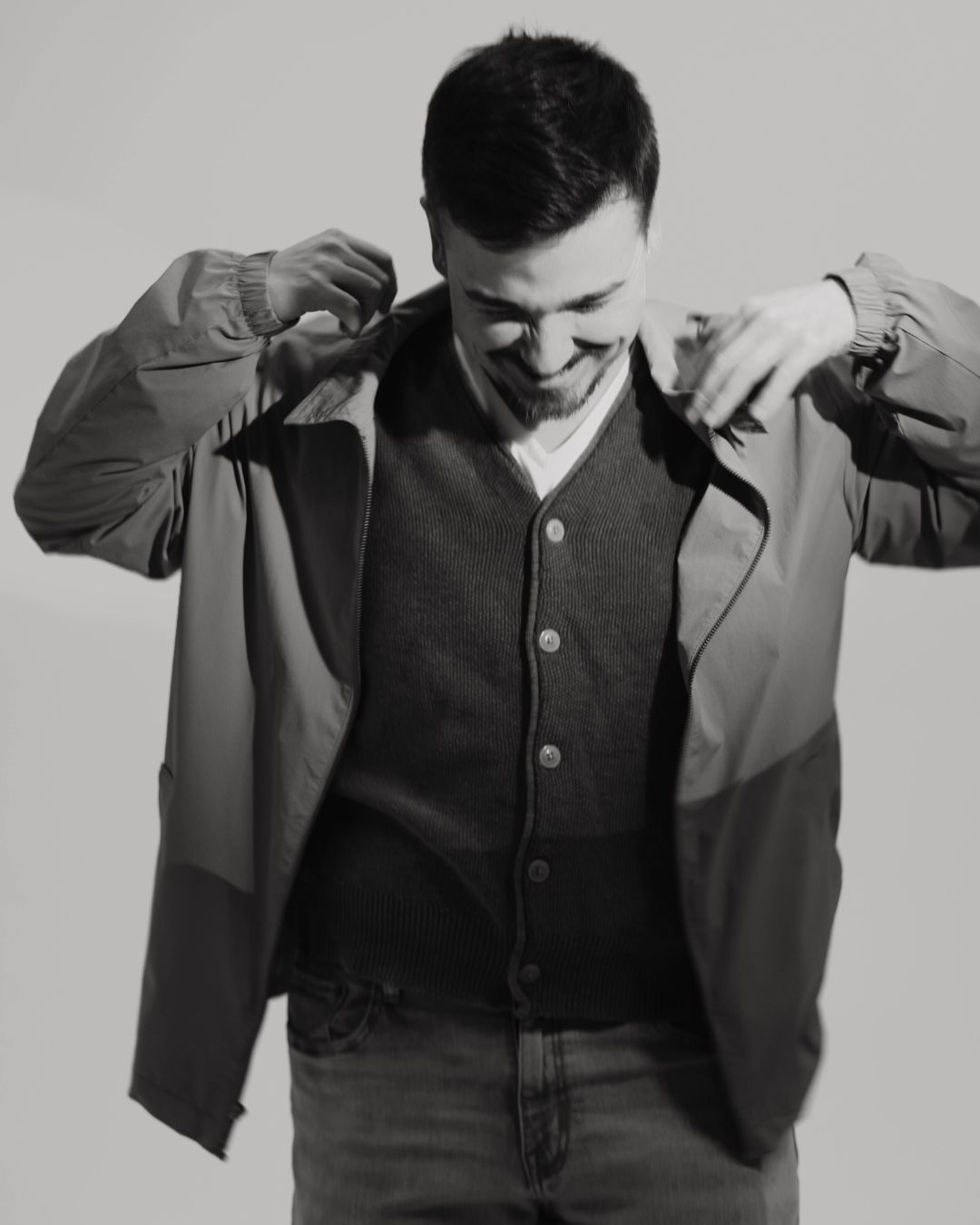 Person adjusting jacket collar, smiling, wearing a dark shirt and light jacket, in black and white.
