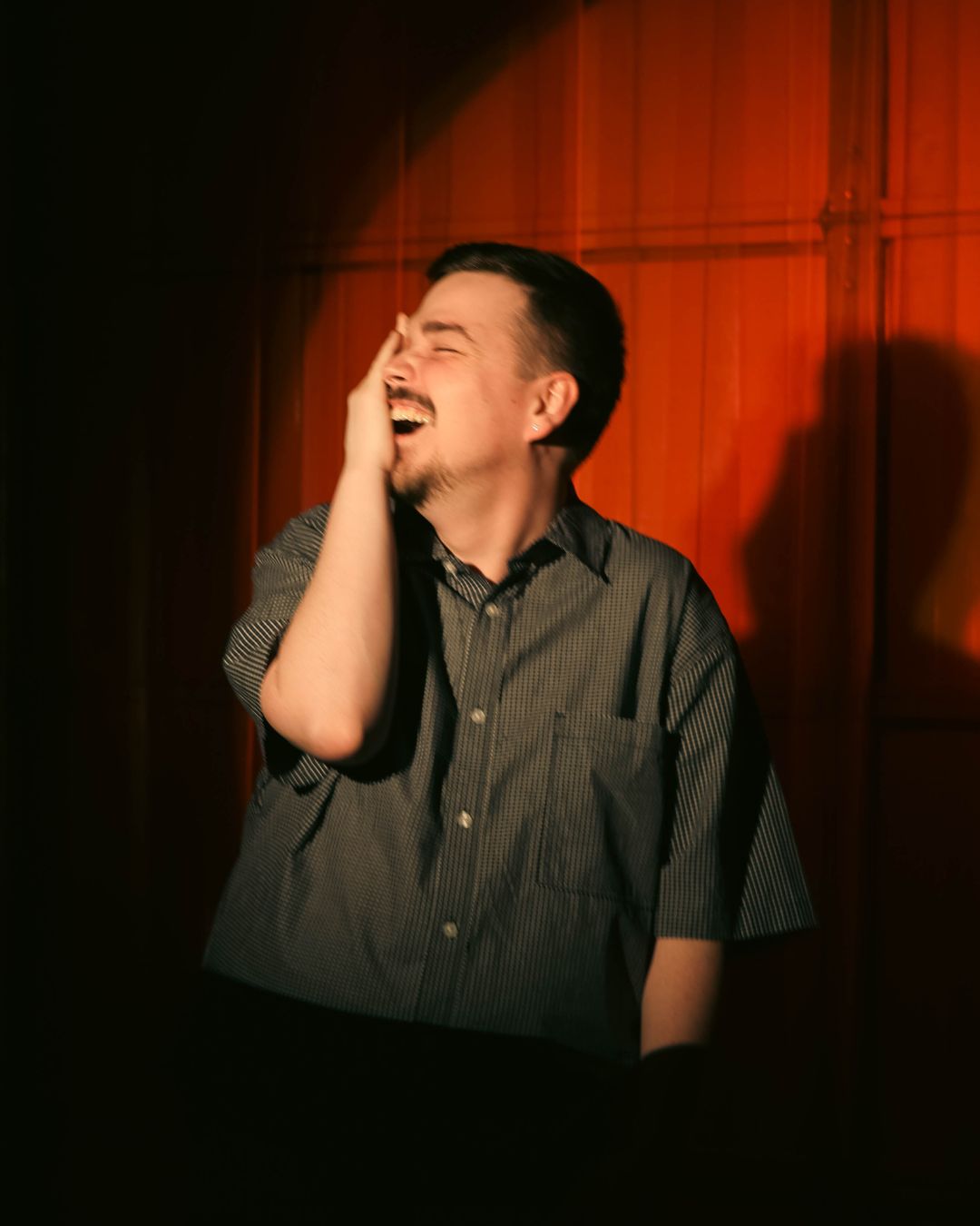 Person laughing with hand on face, standing in front of red curtain, wearing a dark shirt.