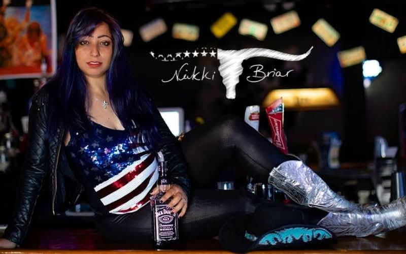 Woman with long dark hair wearing a shiny top and leather jacket, leaning on a bar counter, with a person performing a high kick behind her.