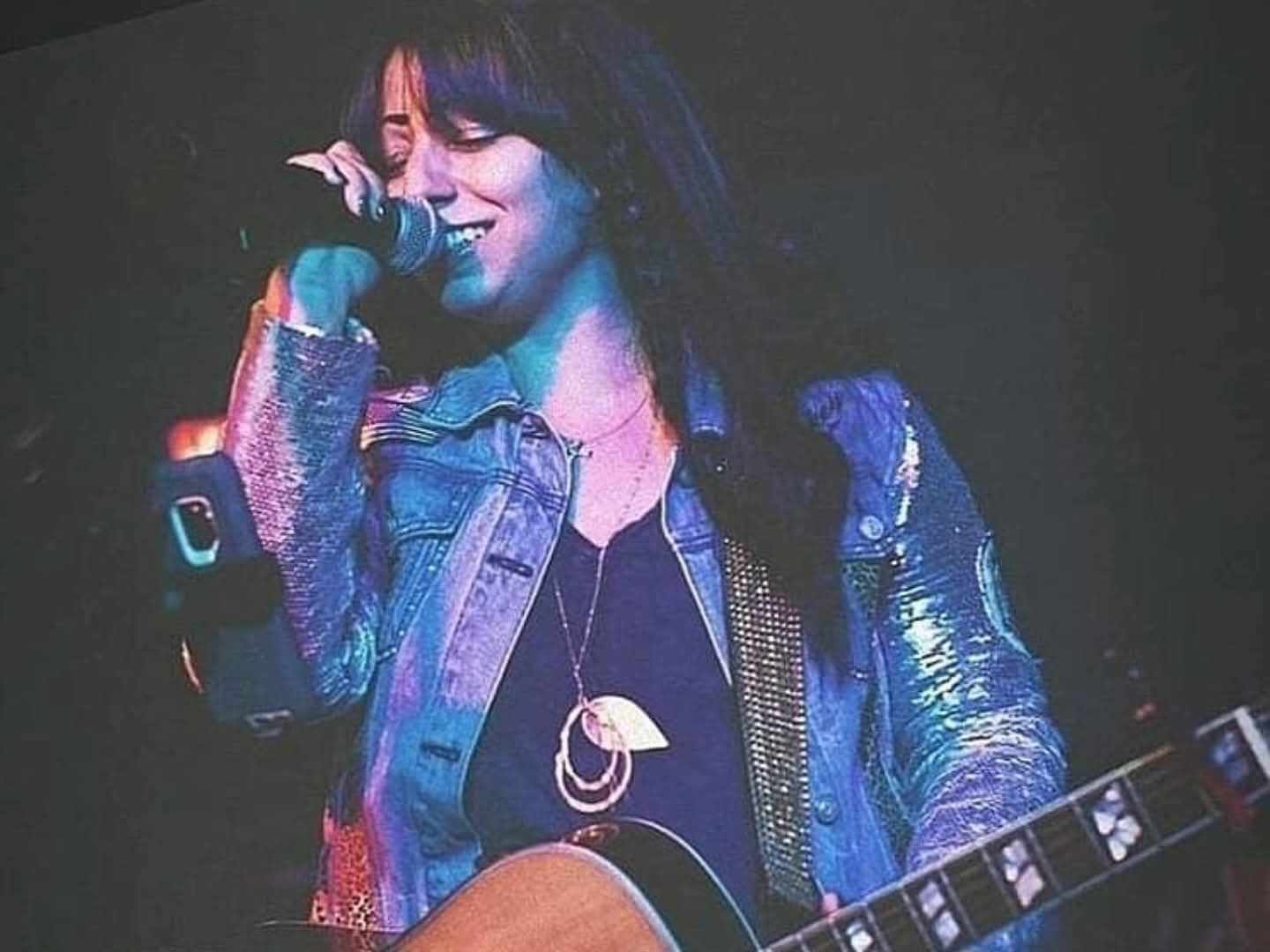 Person singing into a microphone and playing guitar on stage, wearing a denim jacket and necklace.