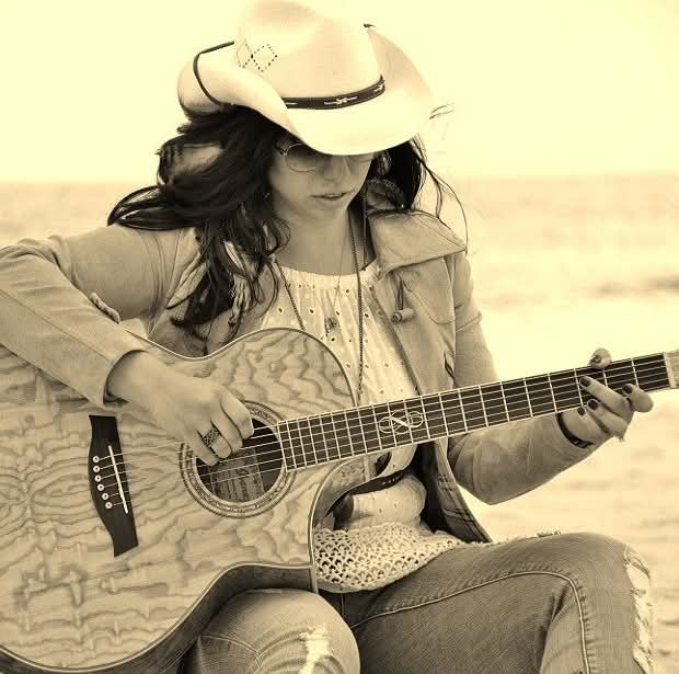 Woman wearing a cowboy hat playing an acoustic guitar outdoors, sitting on a rock or similar surface.