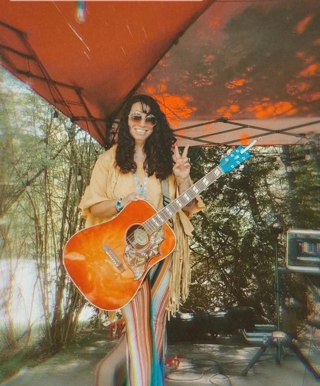 Woman with curly hair and sunglasses holding an orange acoustic guitar outdoors under a canopy, smiling and making a peace sign.