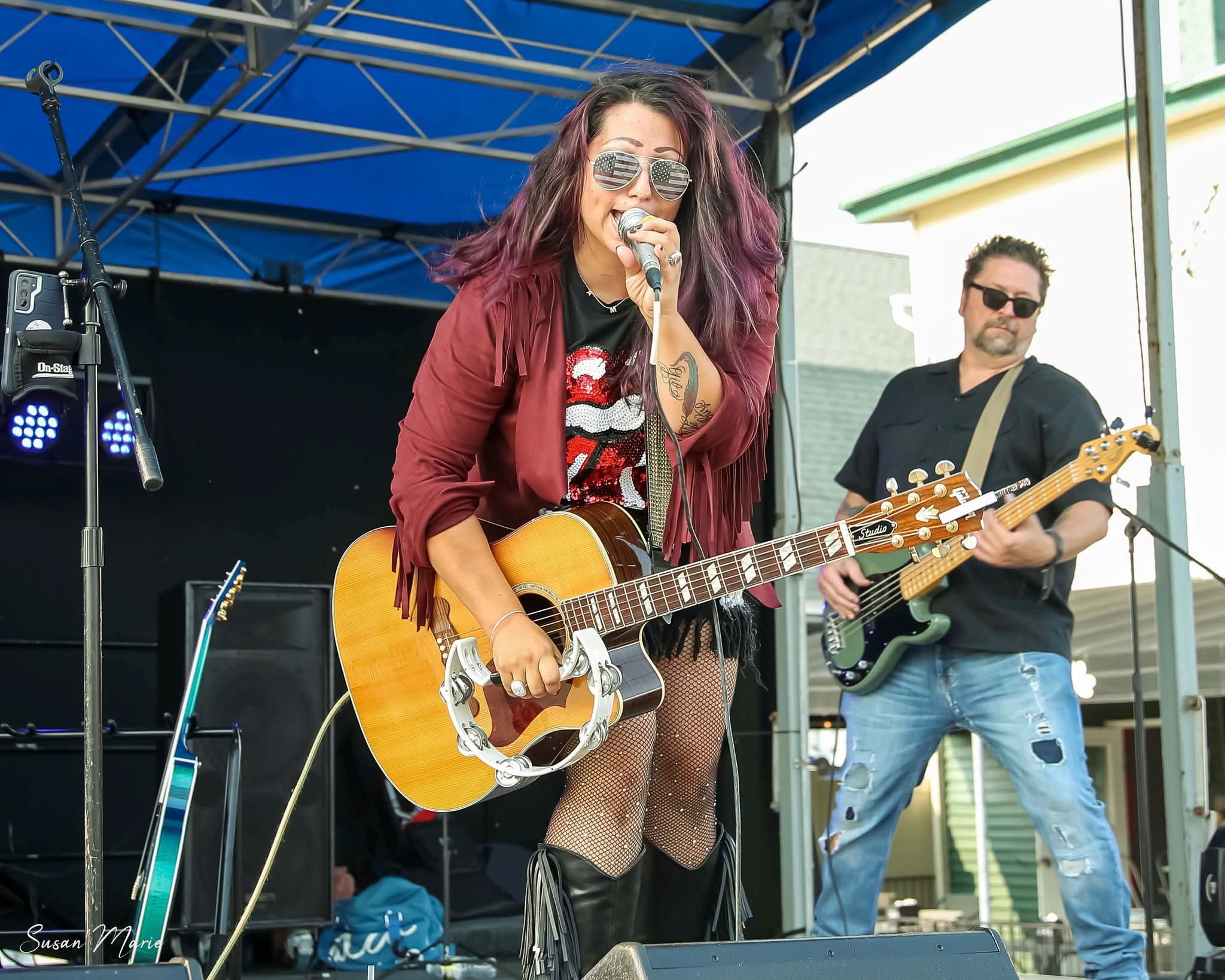 Female musician with long hair, sunglasses, red jacket, playing guitar and singing on stage, male guitarist in background, outdoor setting.