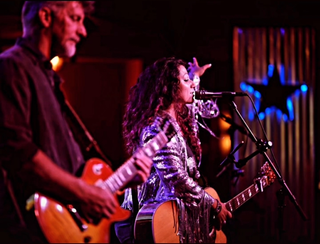 Two musicians perform on stage with guitars, one singing into a microphone, colorful background with star-shaped decoration.
