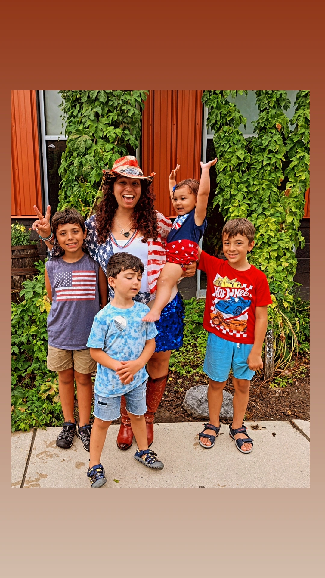 Woman with four children outdoors, some children wearing patriotic clothing, standing in front of a building with greenery.