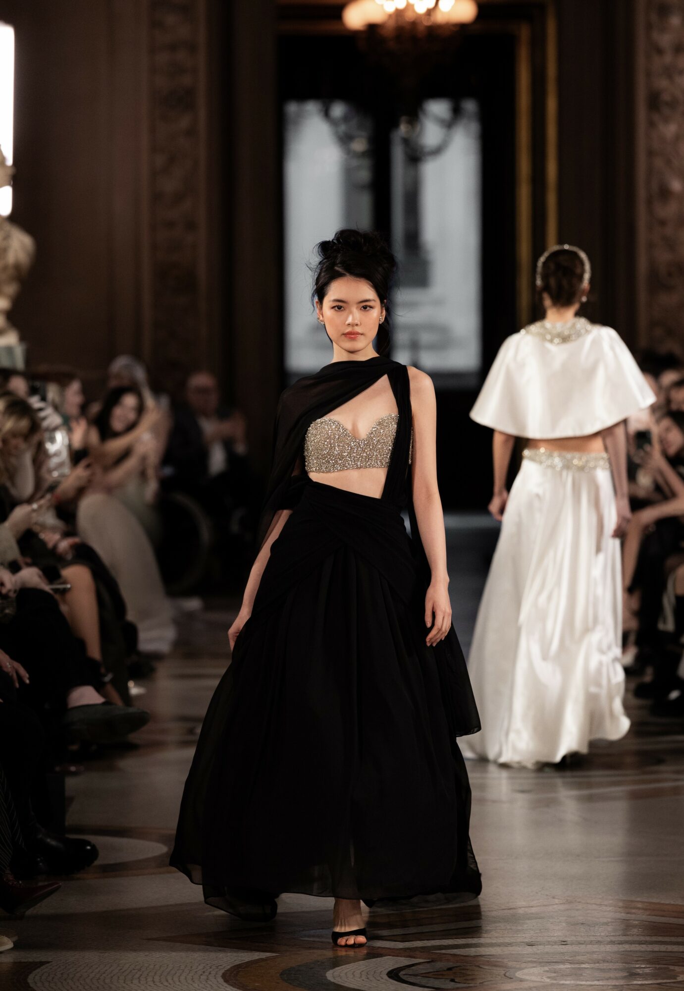Fashion model walking runway in black dress with sheer top, audience seated on sides, ornate room background.