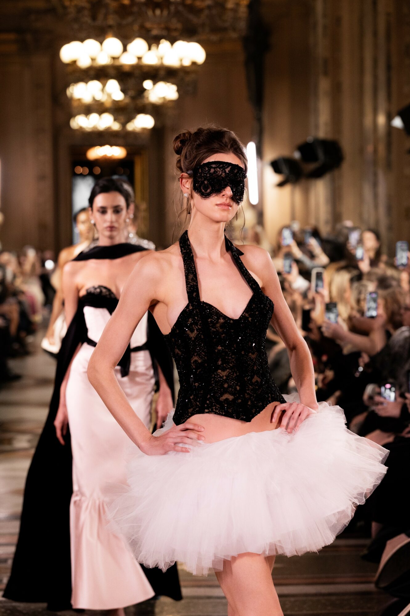 Woman in black corset and white tutu with mask, standing in ornate room with chandeliers and seated audience.