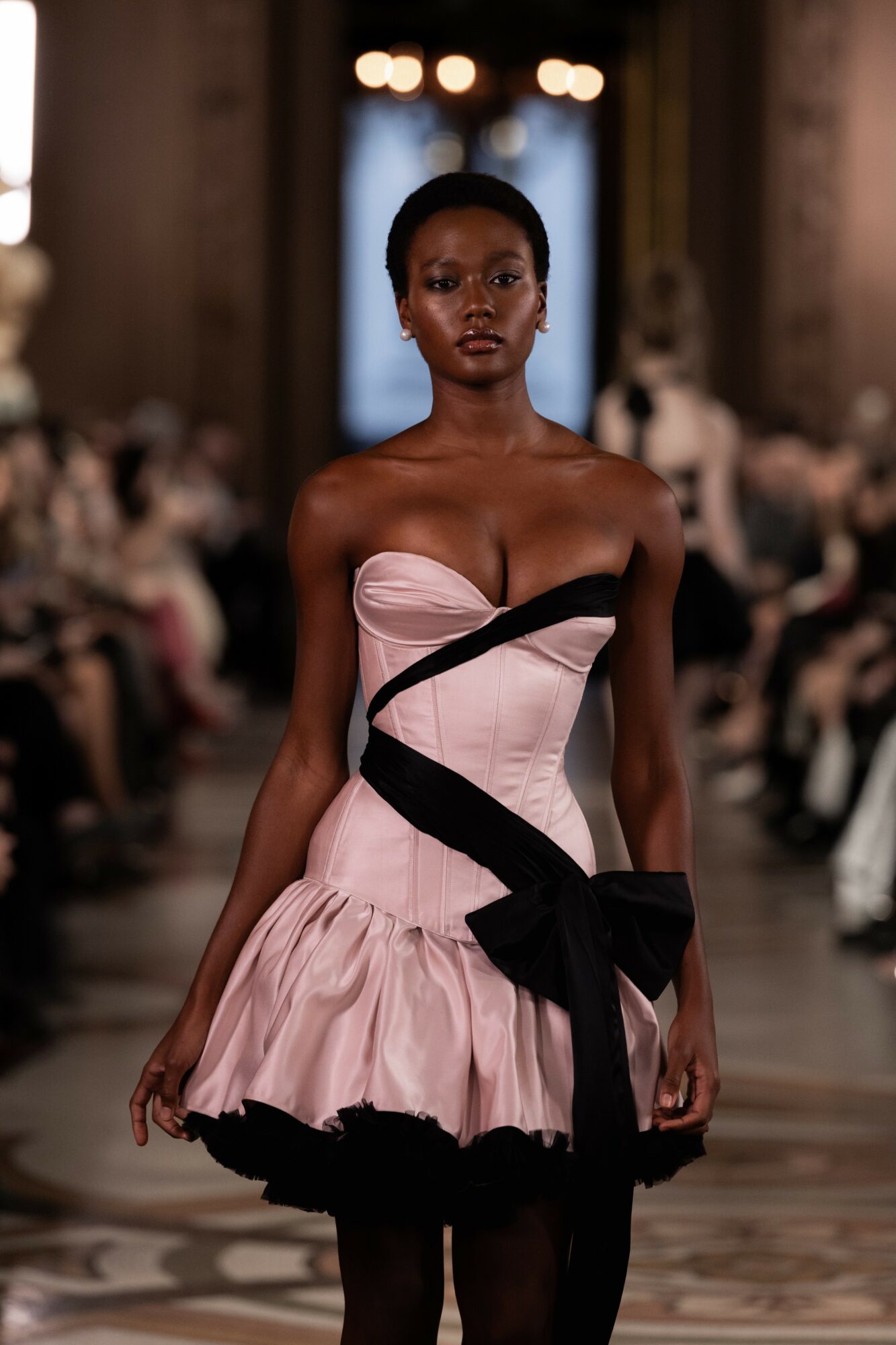 Model walking down runway in pink dress with black accents, audience seated on both sides, ornate interior background.