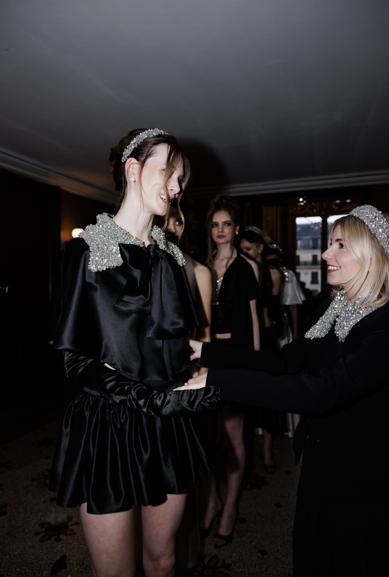 Two women in formal attire holding hands and smiling, with other women in the background, indoors with dark lighting.