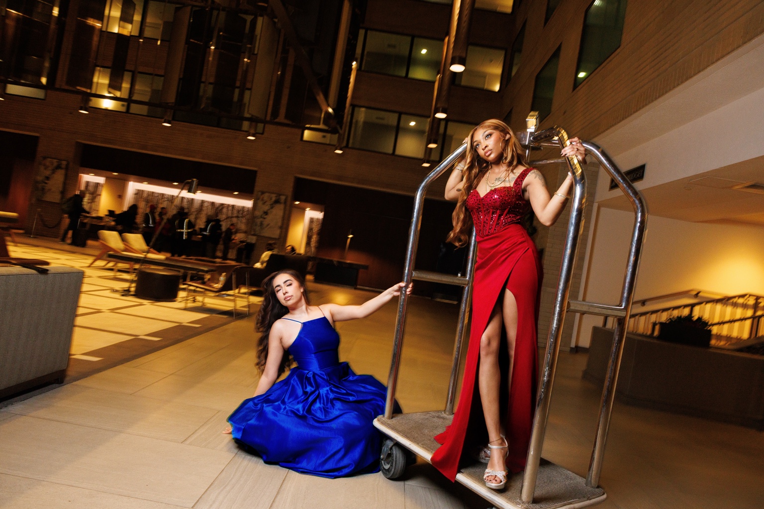 Two women in elegant dresses pose on a hotel lobby luggage cart, one sitting and one standing, in a modern interior.
