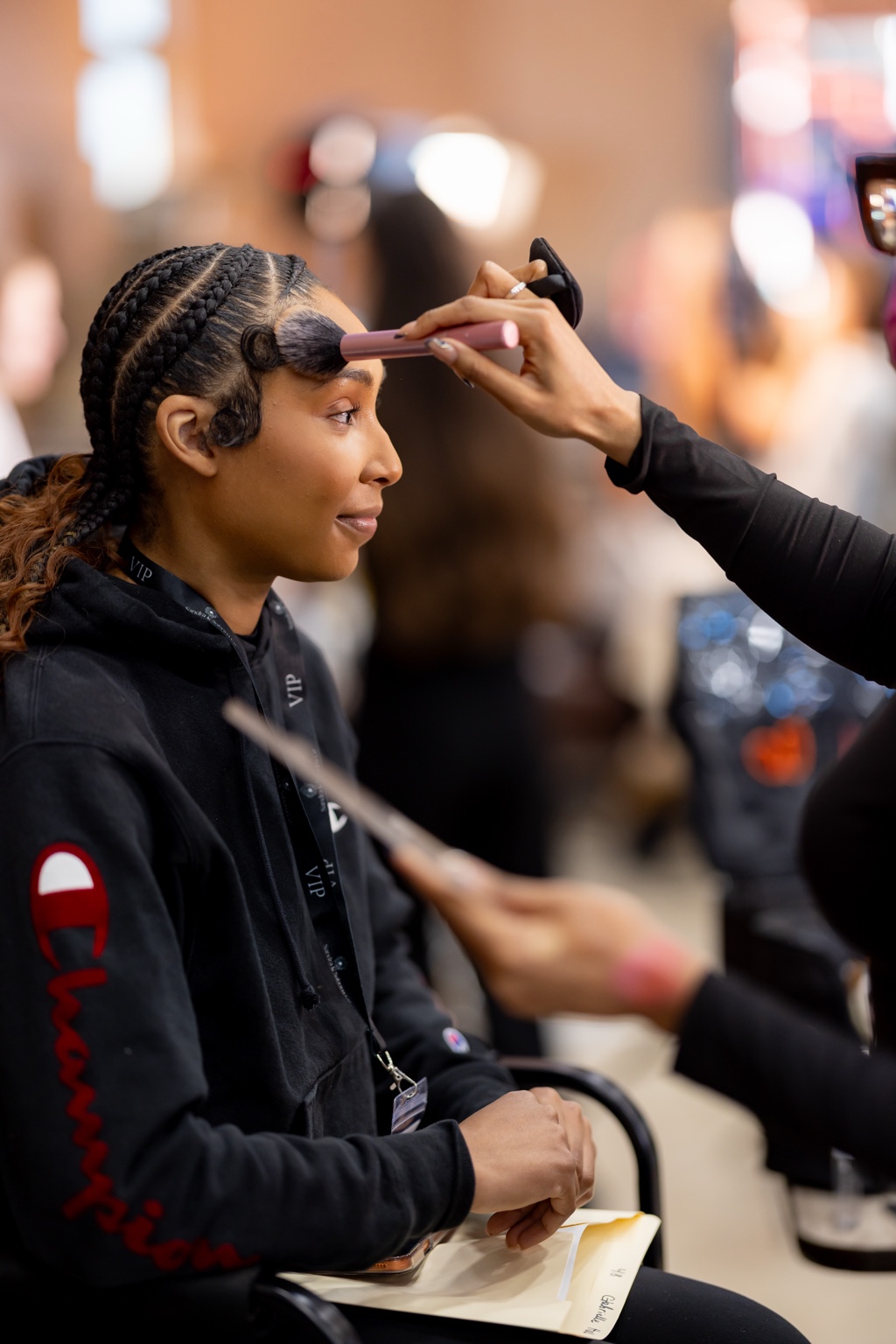 A young girl with braided hair is getting her makeup done by a person in a busy setting.