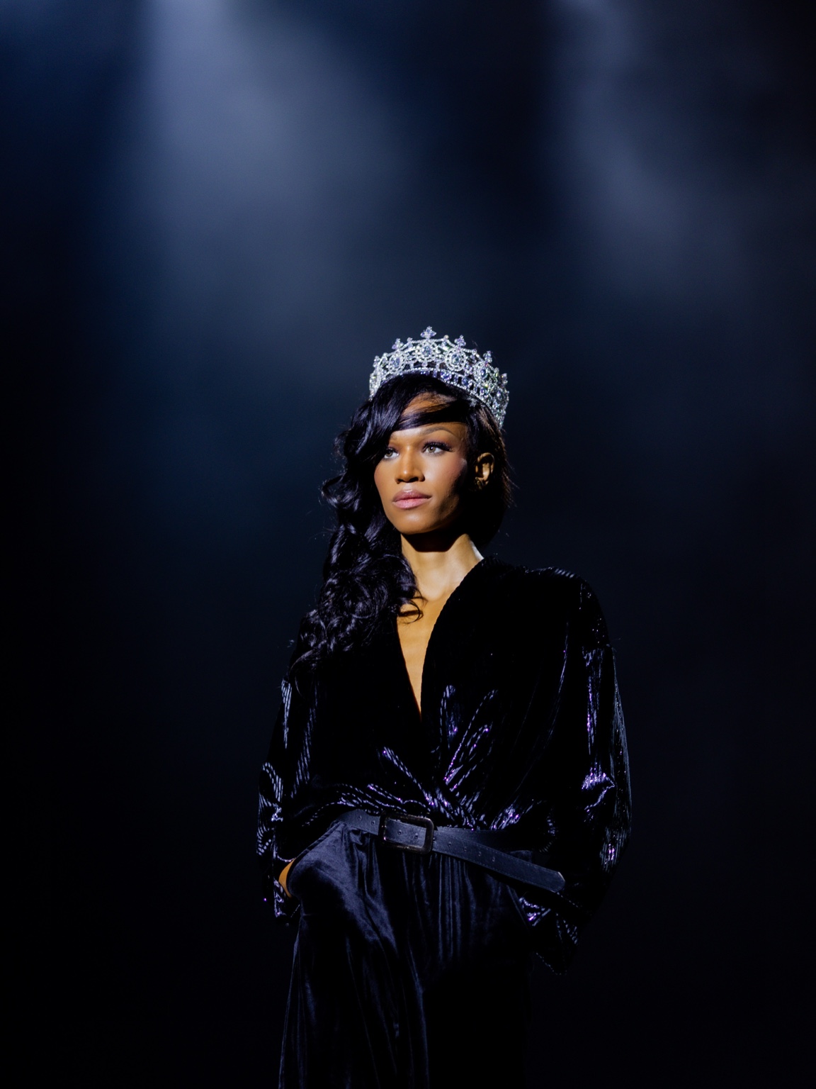 Woman wearing a crown and black velvet dress, standing in a dark setting with spotlight, serious expression.