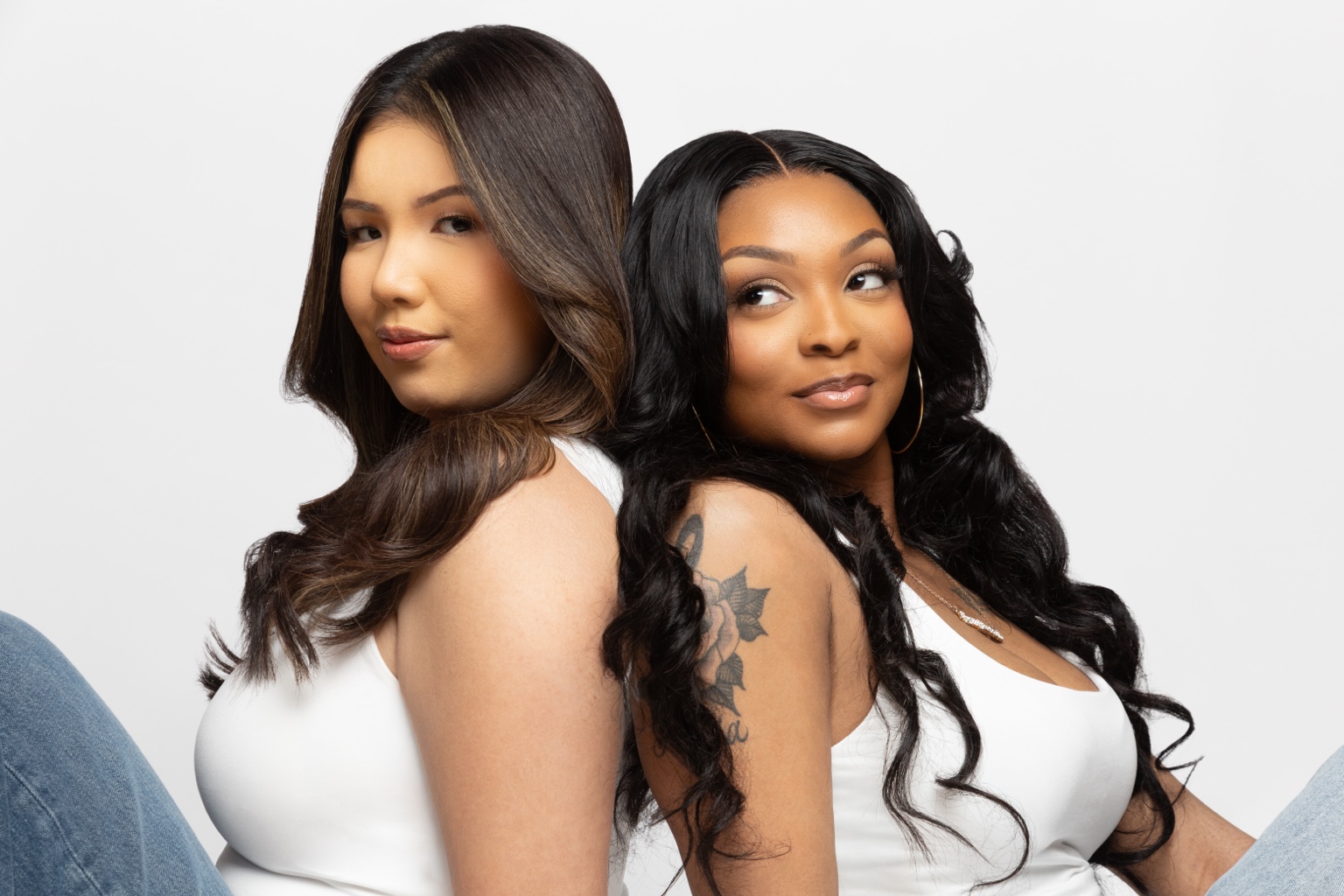 Two women sit close together, one with long dark hair and the other with long wavy black hair, both wearing white tops.