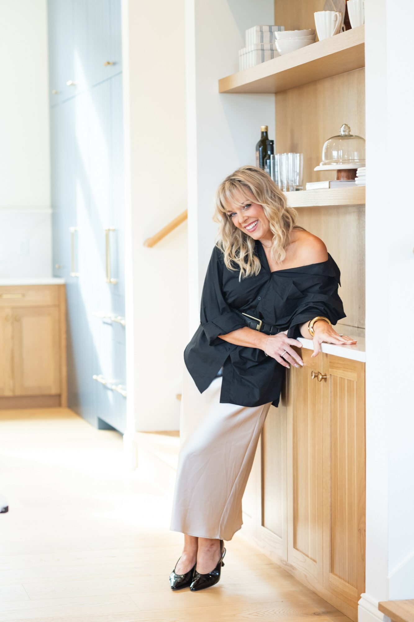 Woman with blonde hair leaning against a kitchen counter, smiling, wearing a black off-shoulder top and beige skirt.
