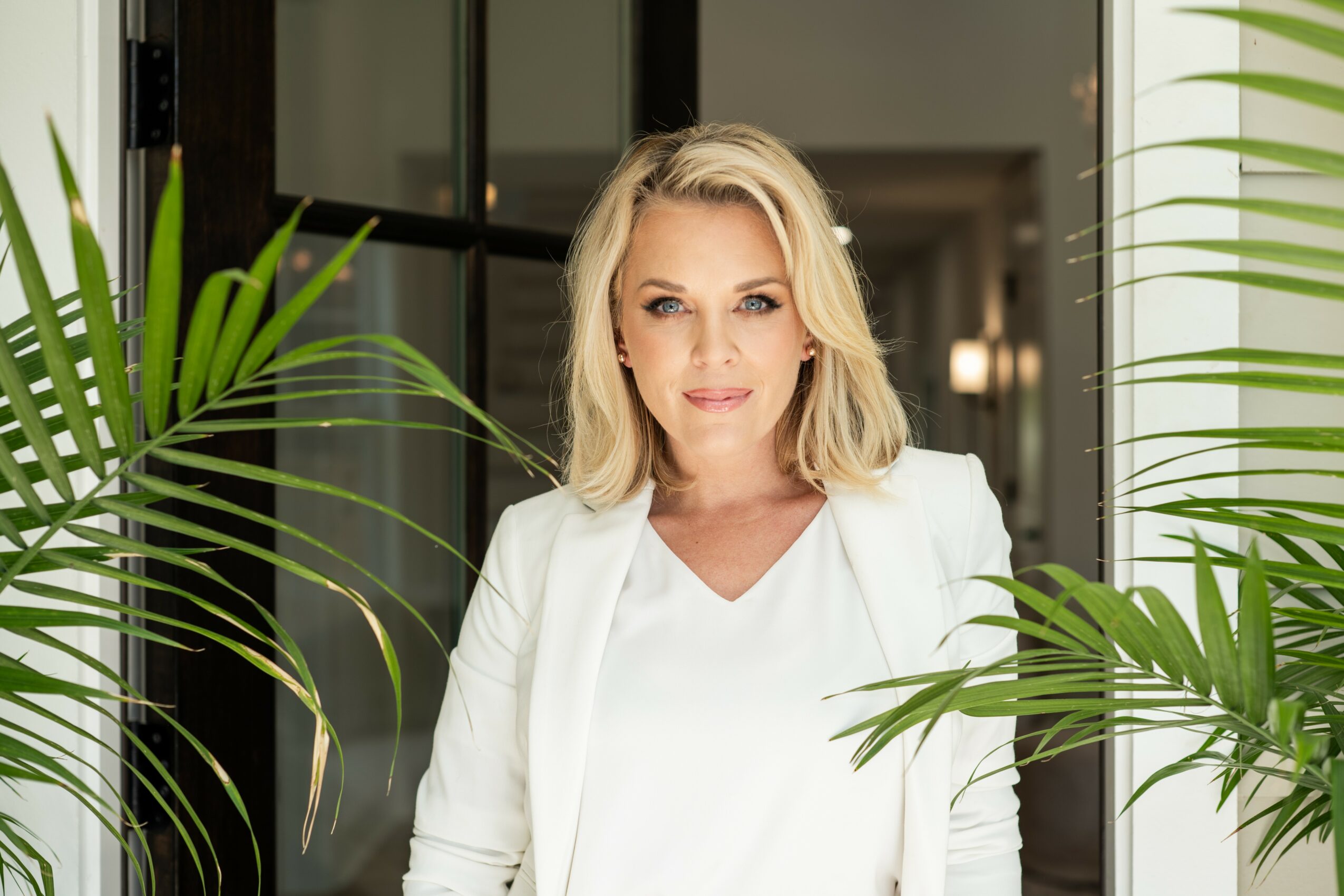 Woman with blonde hair in a white blazer and top, standing indoors with green plants on either side.