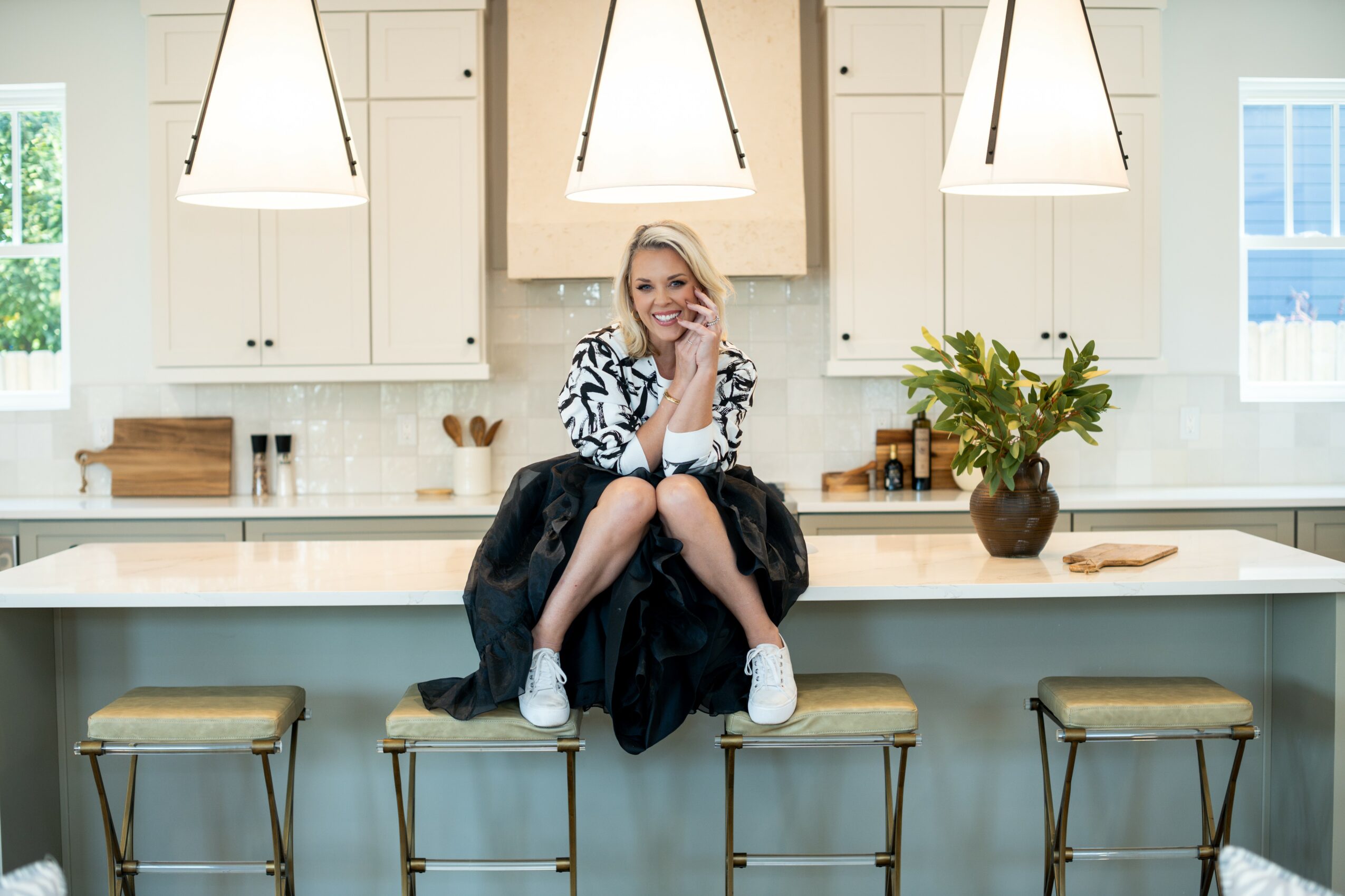 Woman sitting on kitchen counter with three pendant lights above, white cabinets, and a potted plant nearby.