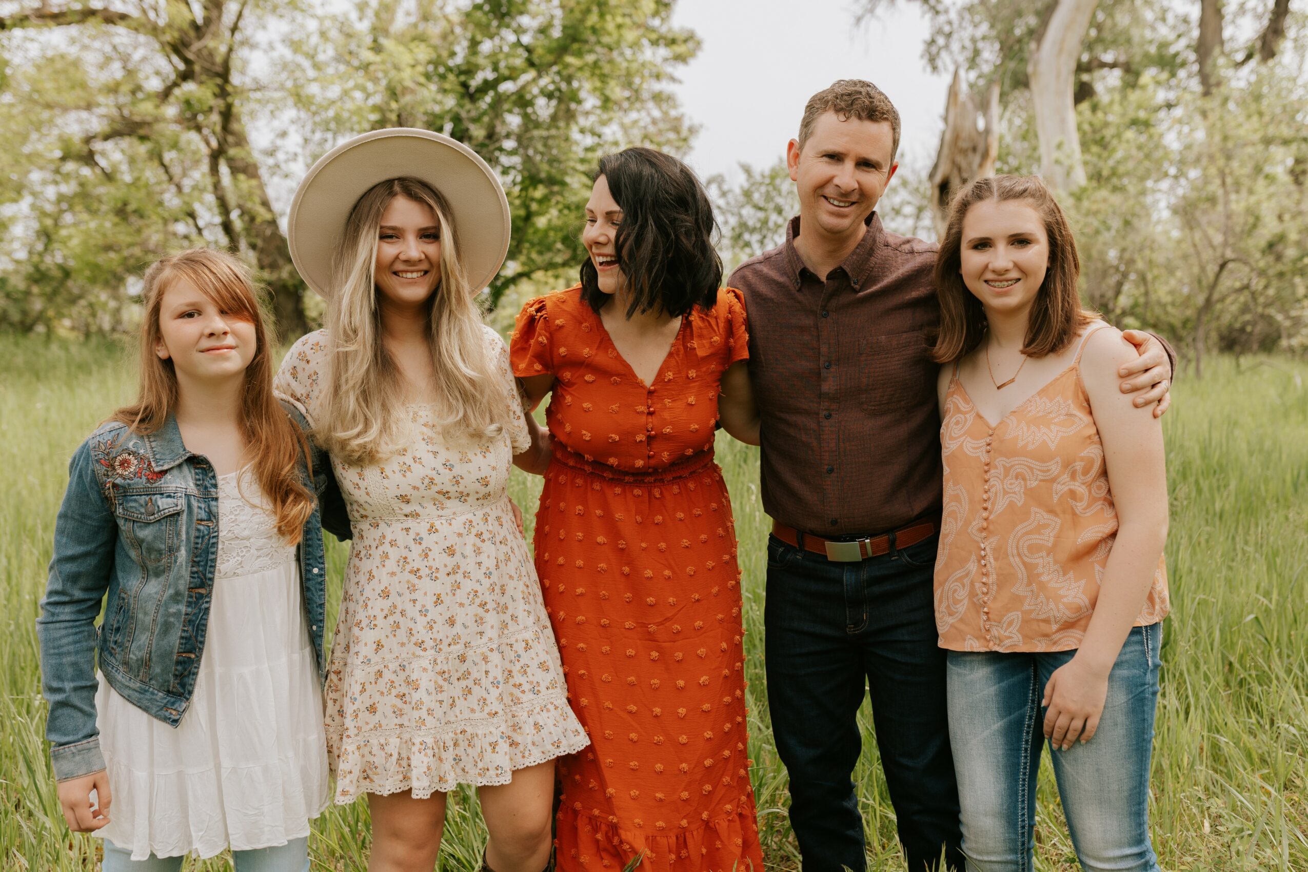 Group of six people standing outdoors in a grassy area with trees, smiling and posing together.