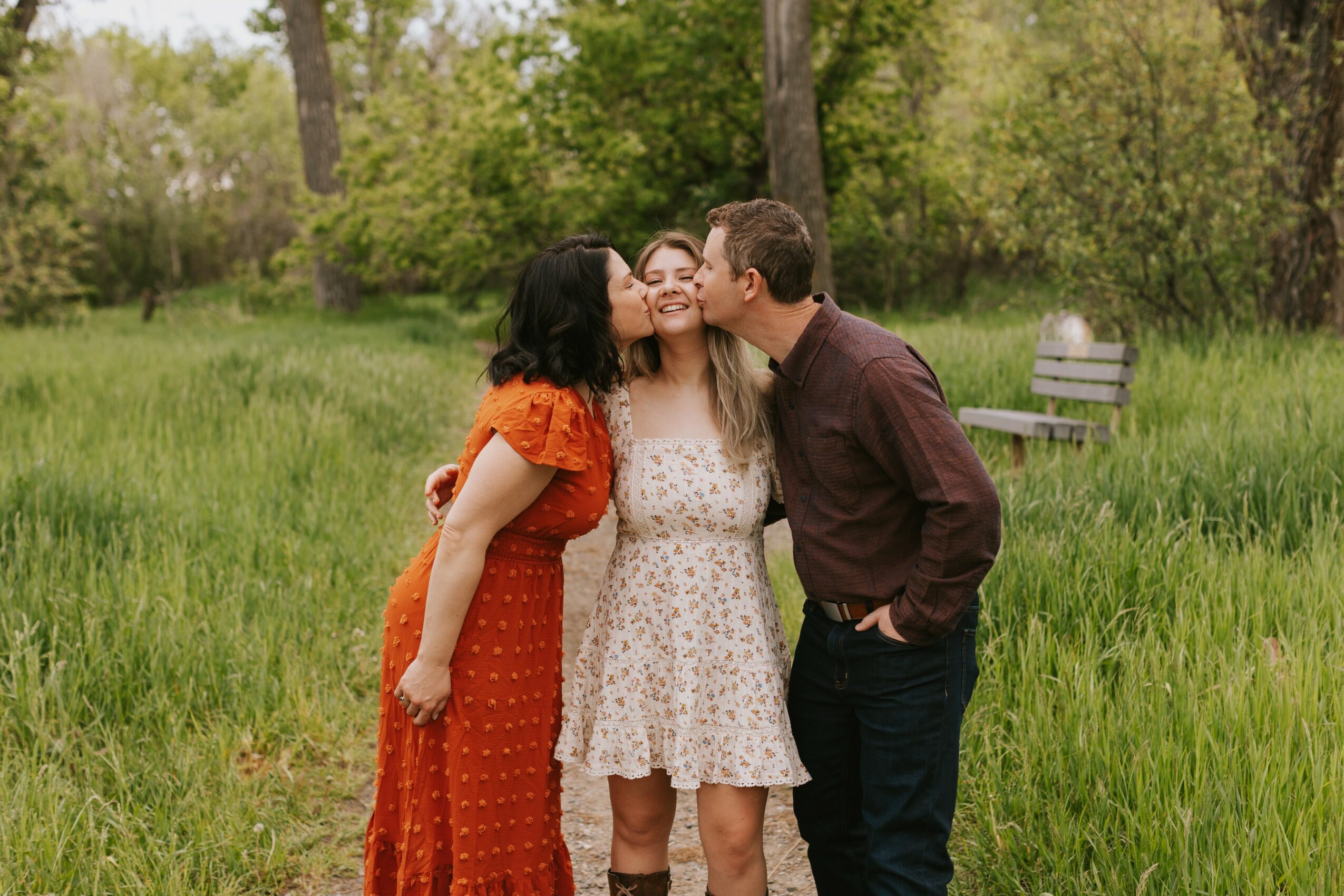 Three people standing close together in a park, smiling and kissing, with trees and a bench in the background.