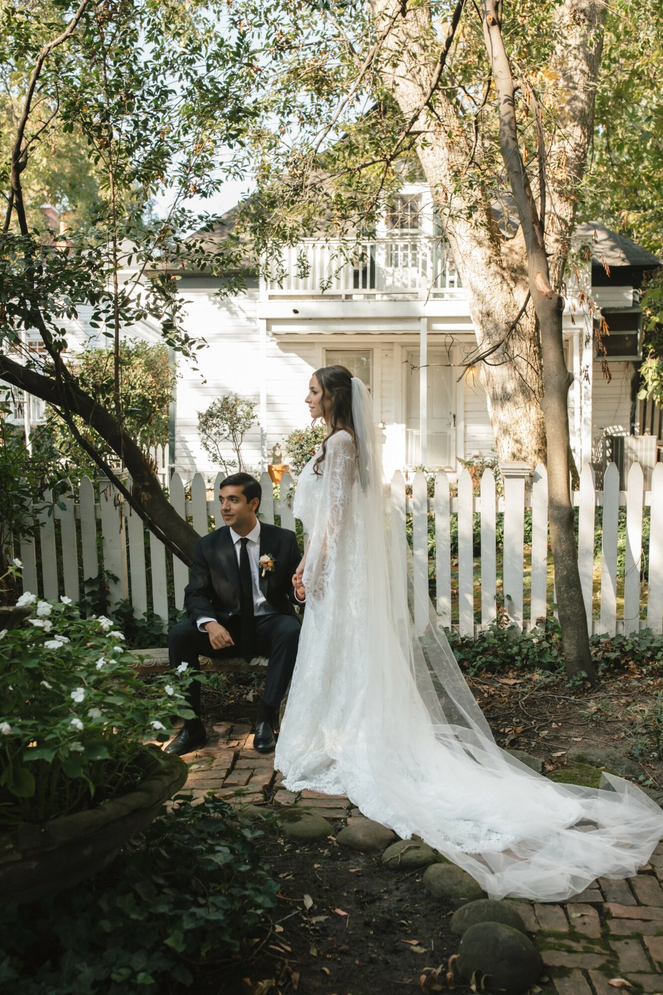 A bride in a long white wedding dress and a groom in a suit sit outdoors near a white picket fence, surrounded by trees and greenery.