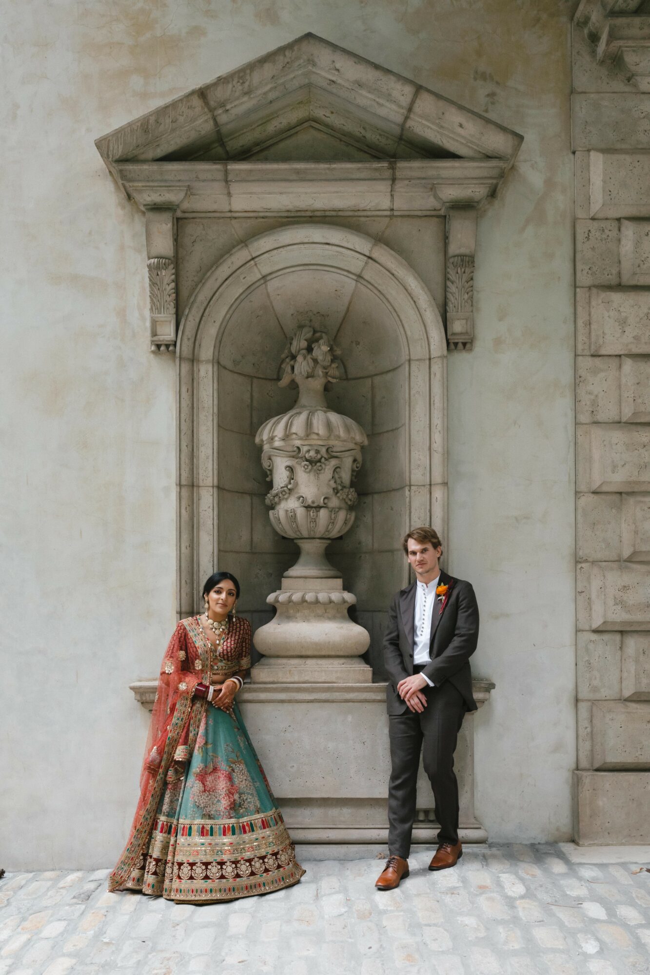 A woman in traditional attire and a man in a suit stand in front of a stone fountain with classical architecture.