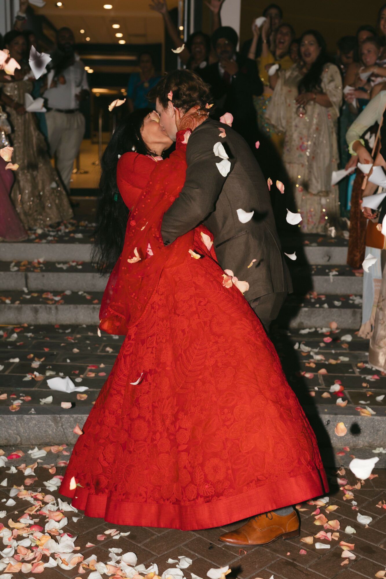 Couple dancing on stairs surrounded by guests, confetti on ground, woman in red gown, man in dark suit, romantic scene