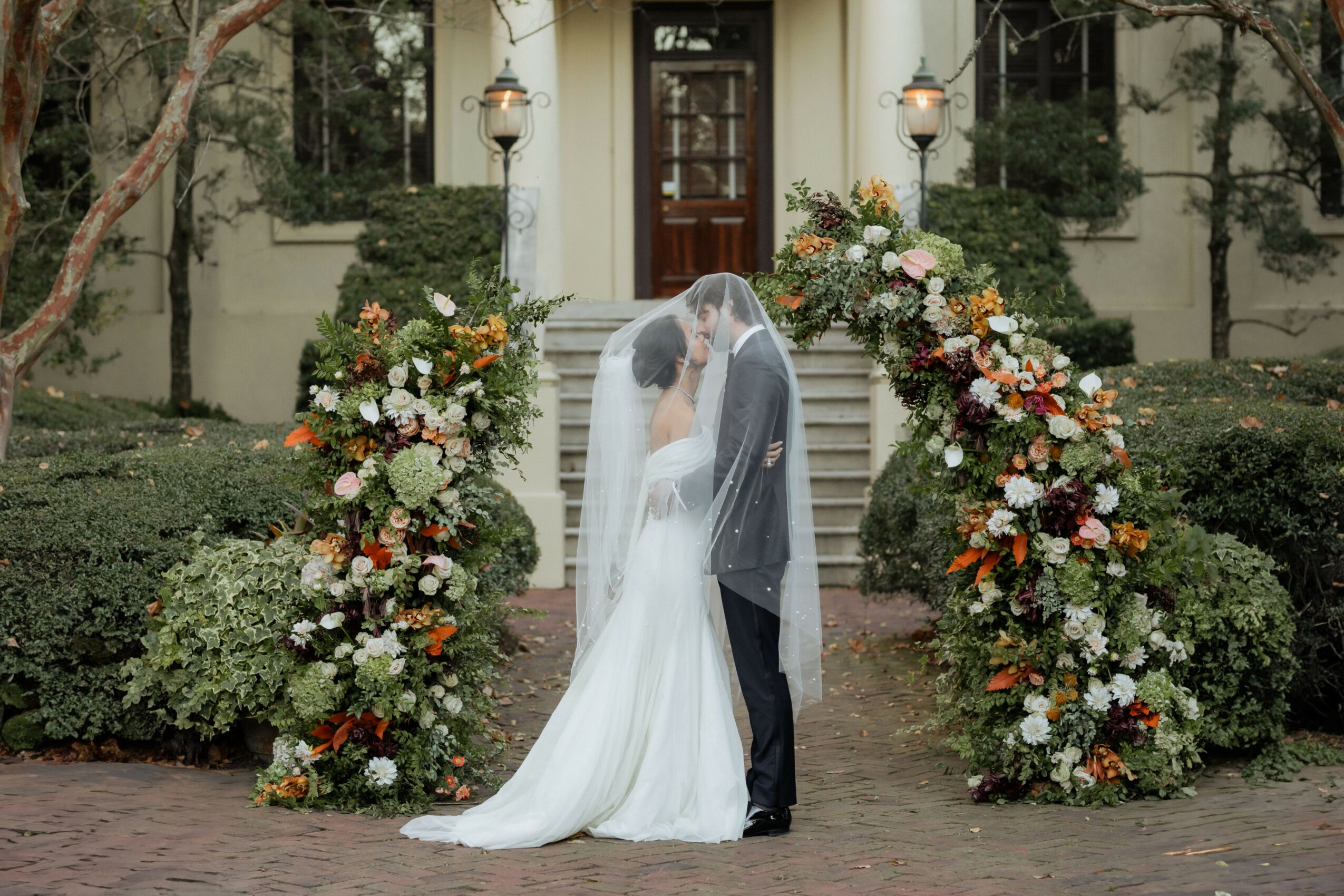 Bride and groom kissing in front of floral arch on steps outside building, with greenery and trees around.