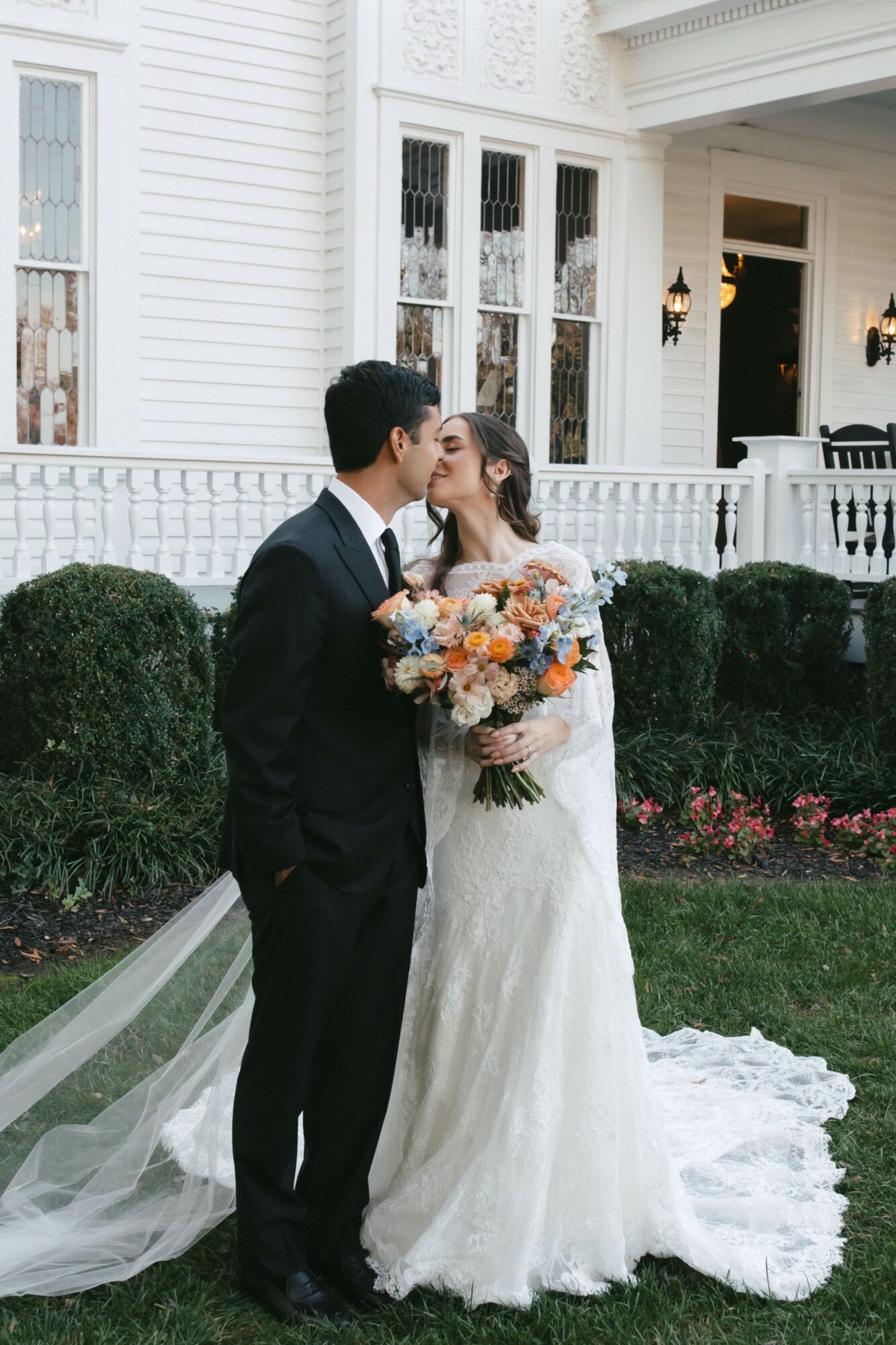 A bride and groom kiss outdoors in front of a white house with large windows and a porch, holding a bouquet.