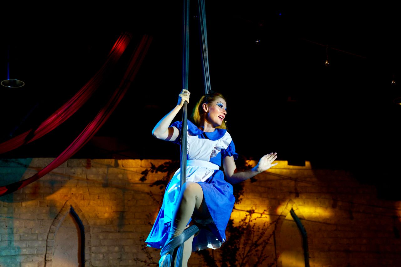 A woman in a blue and white costume performs on a swing, with a dark background and stage lighting.