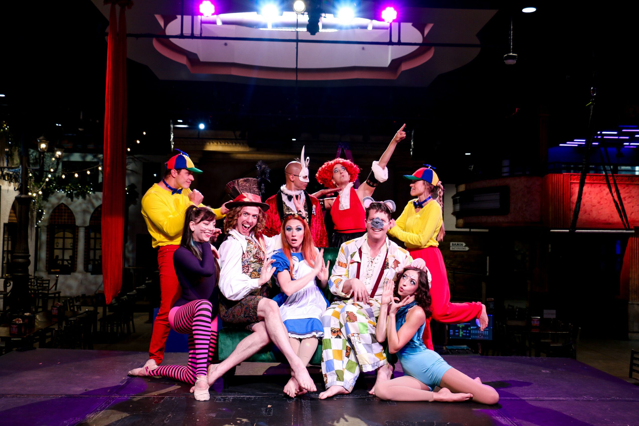 Group of children and adults performing on stage with colorful costumes and props, under stage lighting.