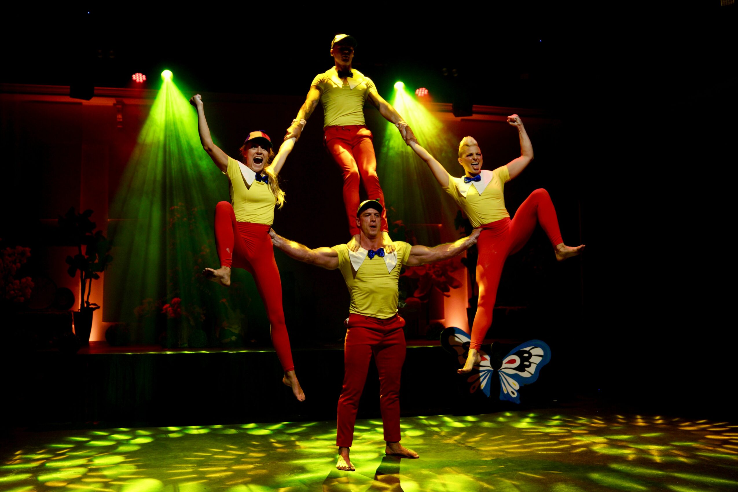 Four performers in yellow tops and red pants perform a dance on stage with green and yellow lighting, one person lifted above others.