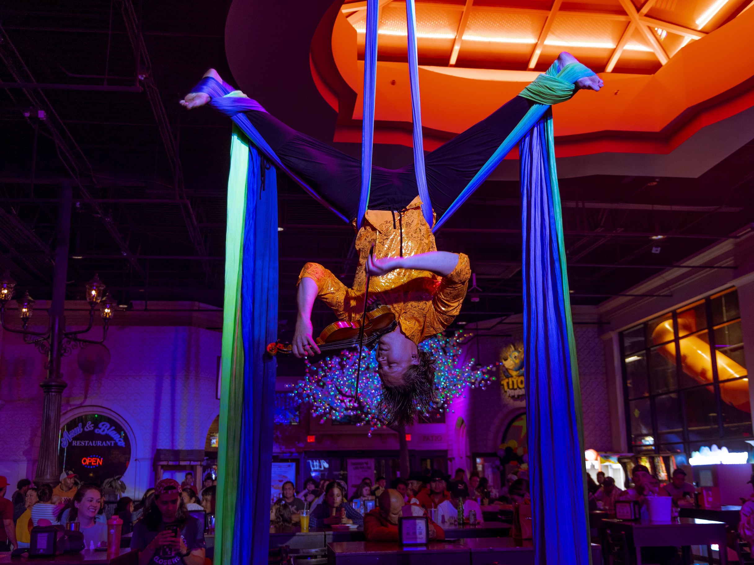 Performer upside down on aerial silk, audience watching in a dimly lit venue with colorful lighting.