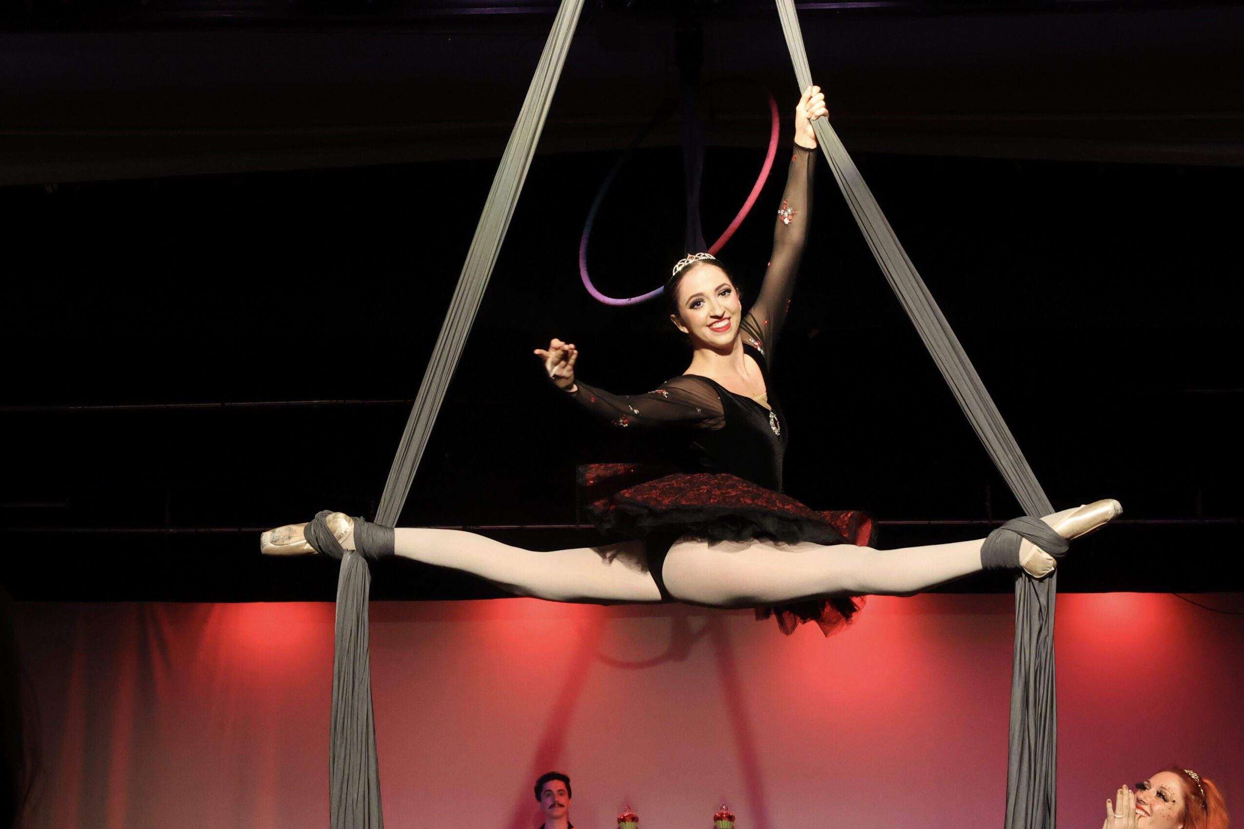 A woman performs an aerial act on a hoop, sitting with legs extended and arms raised, against a dark background with red lighting.