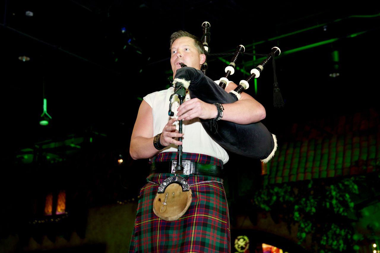 Person playing bagpipes on stage with green lighting in background, wearing a plaid skirt and white top.