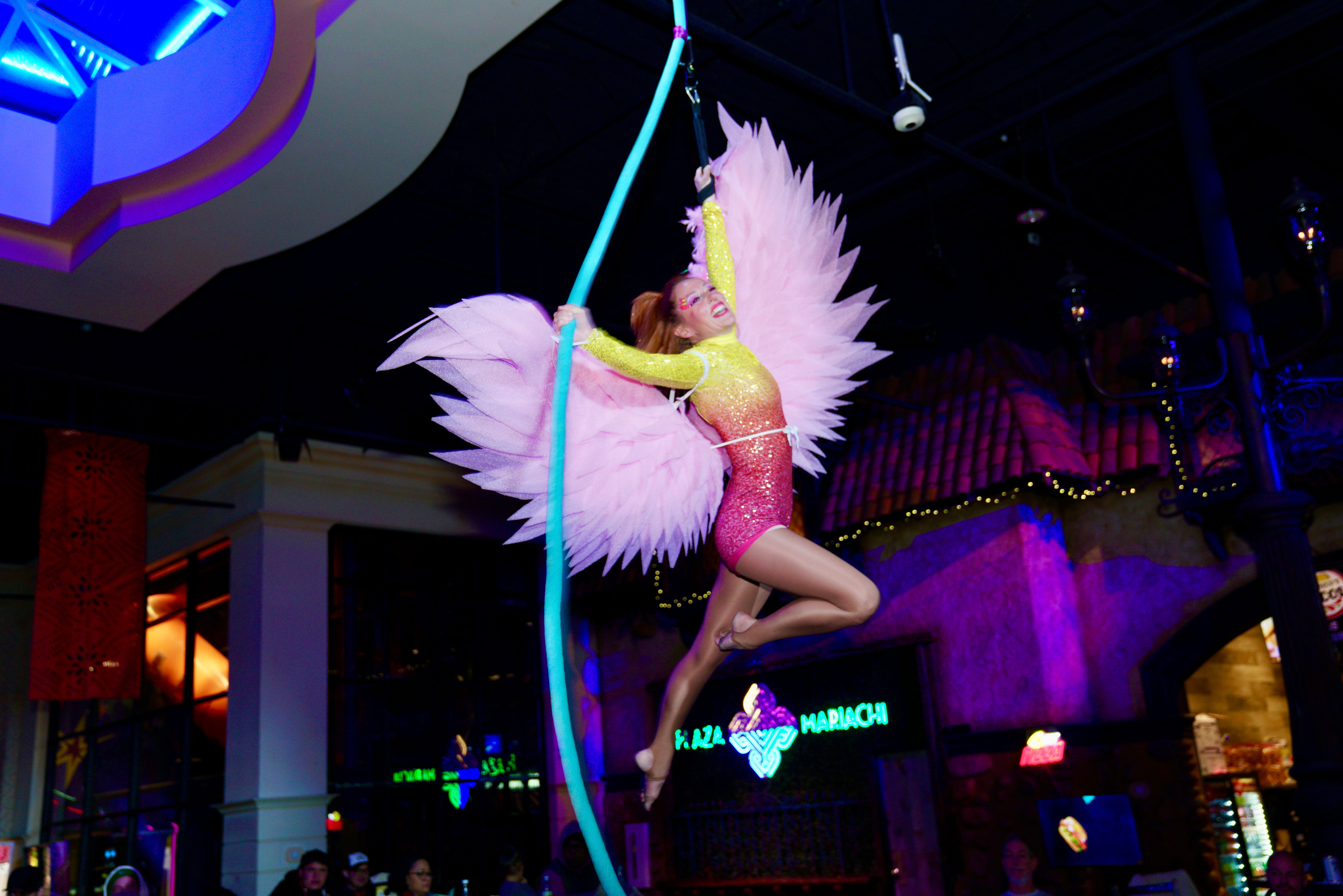 Performer in pink costume with large white wings, hanging from a pole in a nightclub setting.