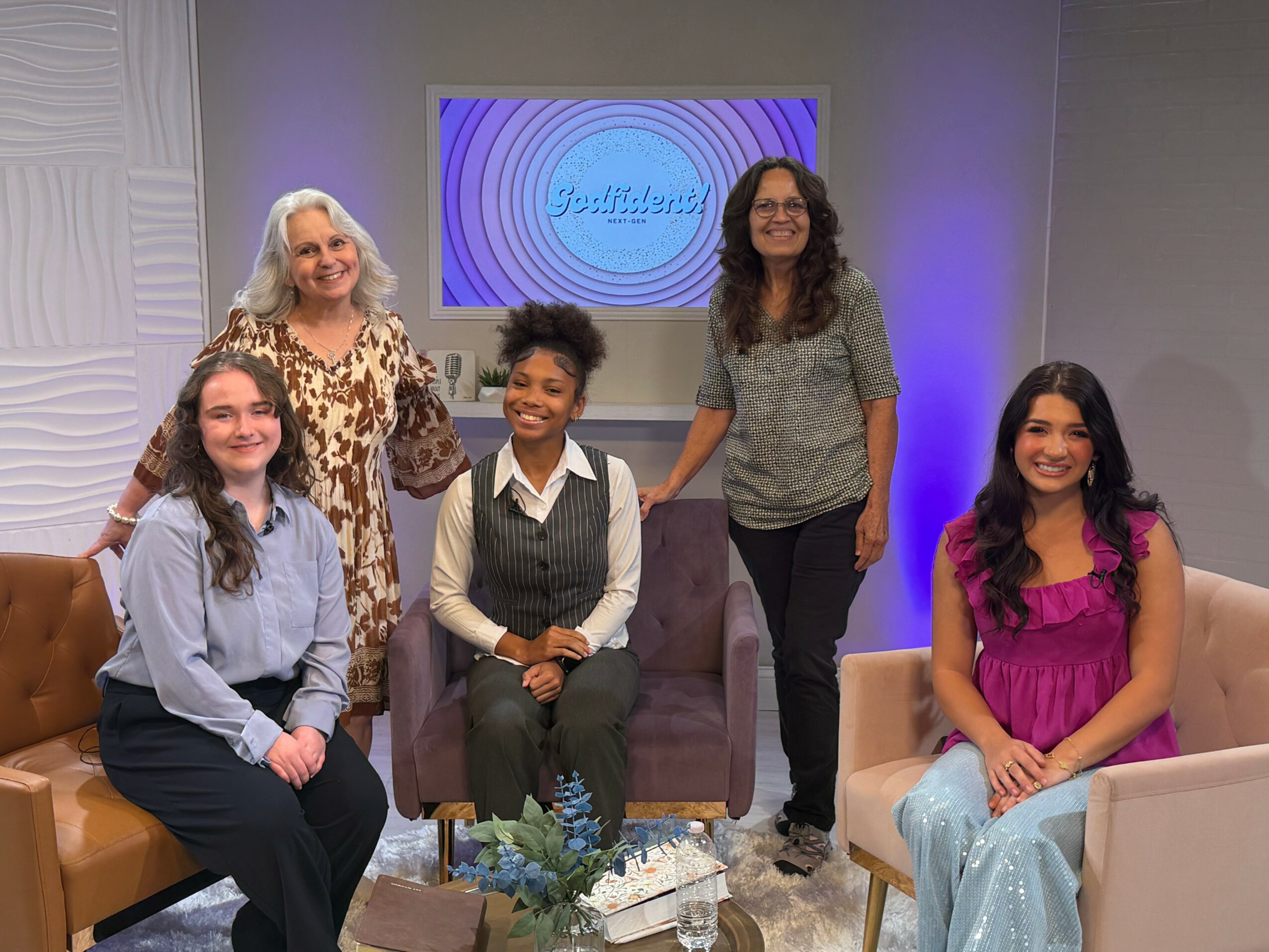 Five women and girls in a studio with a TV screen displaying a logo, sitting and standing around chairs, smiling.