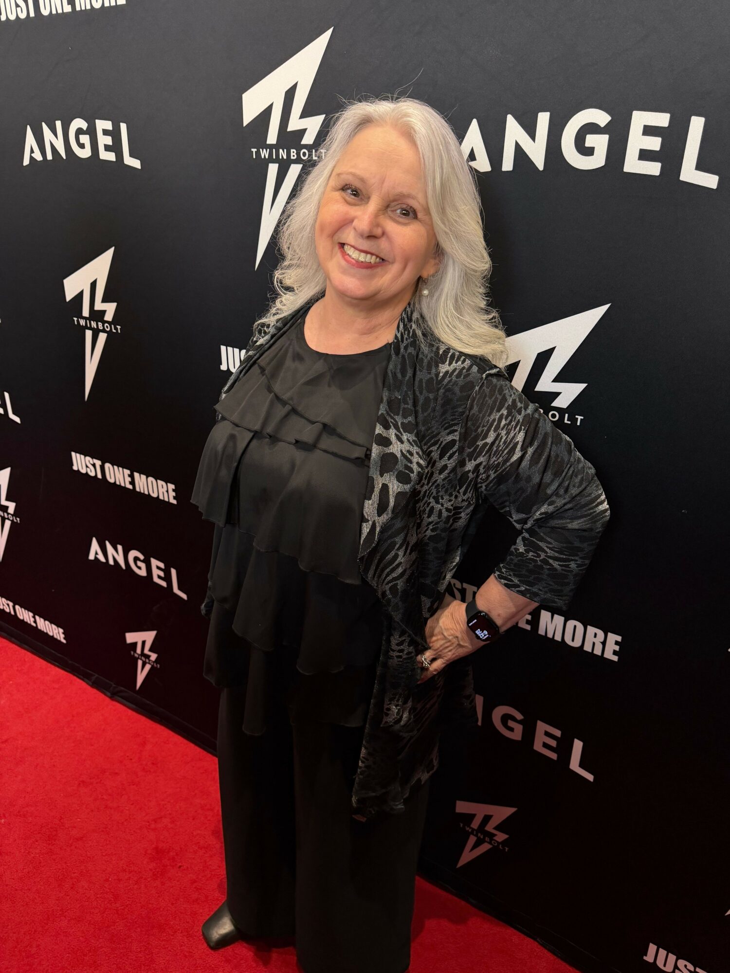 Woman with blonde hair smiling, standing on red carpet, black background with white text and logos, wearing black outfit.